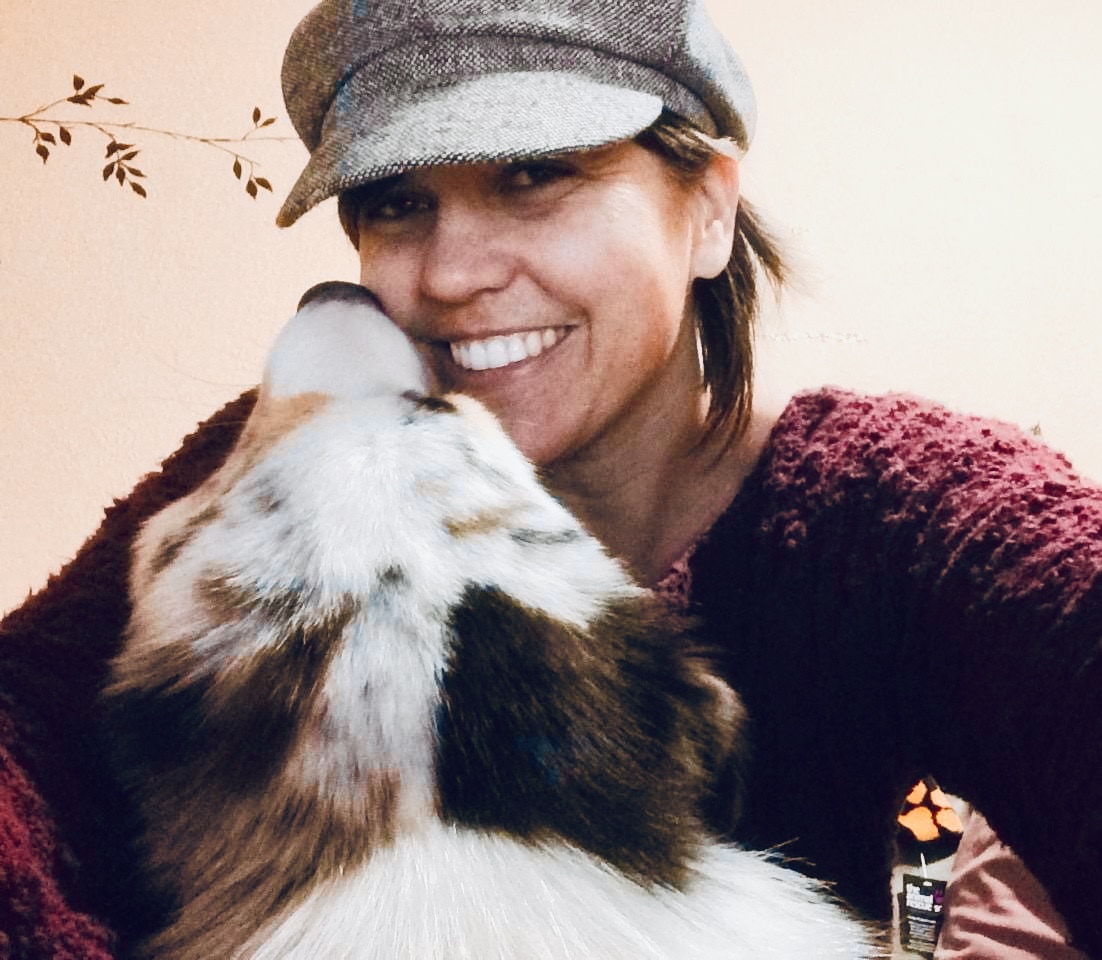 Woman wearing a gray cap smiling as a dog licks her face, indoors with a light background.
