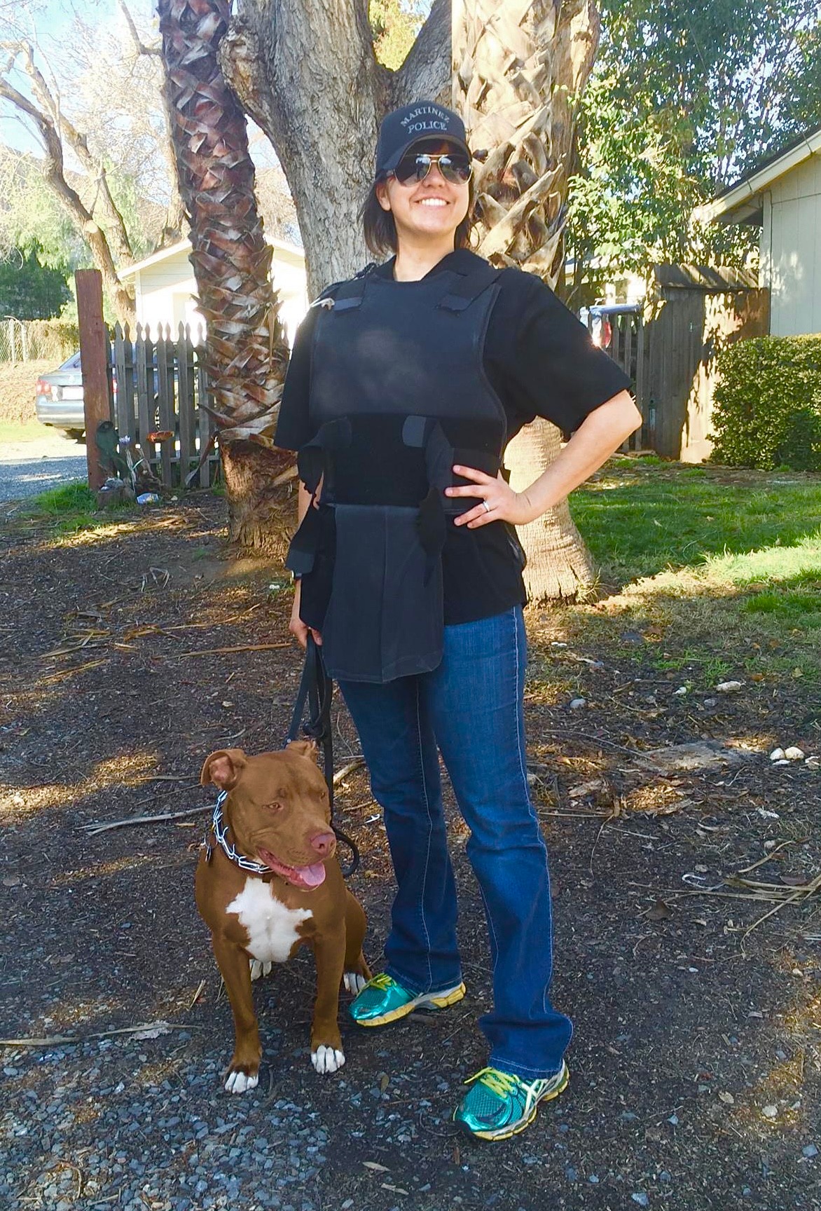 Woman standing outdoors with a brown dog on a leash, smiling, wearing sunglasses, a black shirt, and jeans.