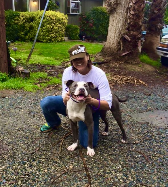 Person crouching outdoors with a happy dog on a leash, smiling, in a green yard with trees and bushes.