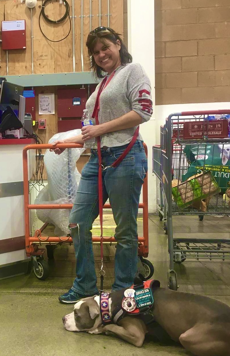 Woman standing in a store aisle with shopping carts, smiling, wearing a gray sweater and jeans, with a dog lying on the floor.