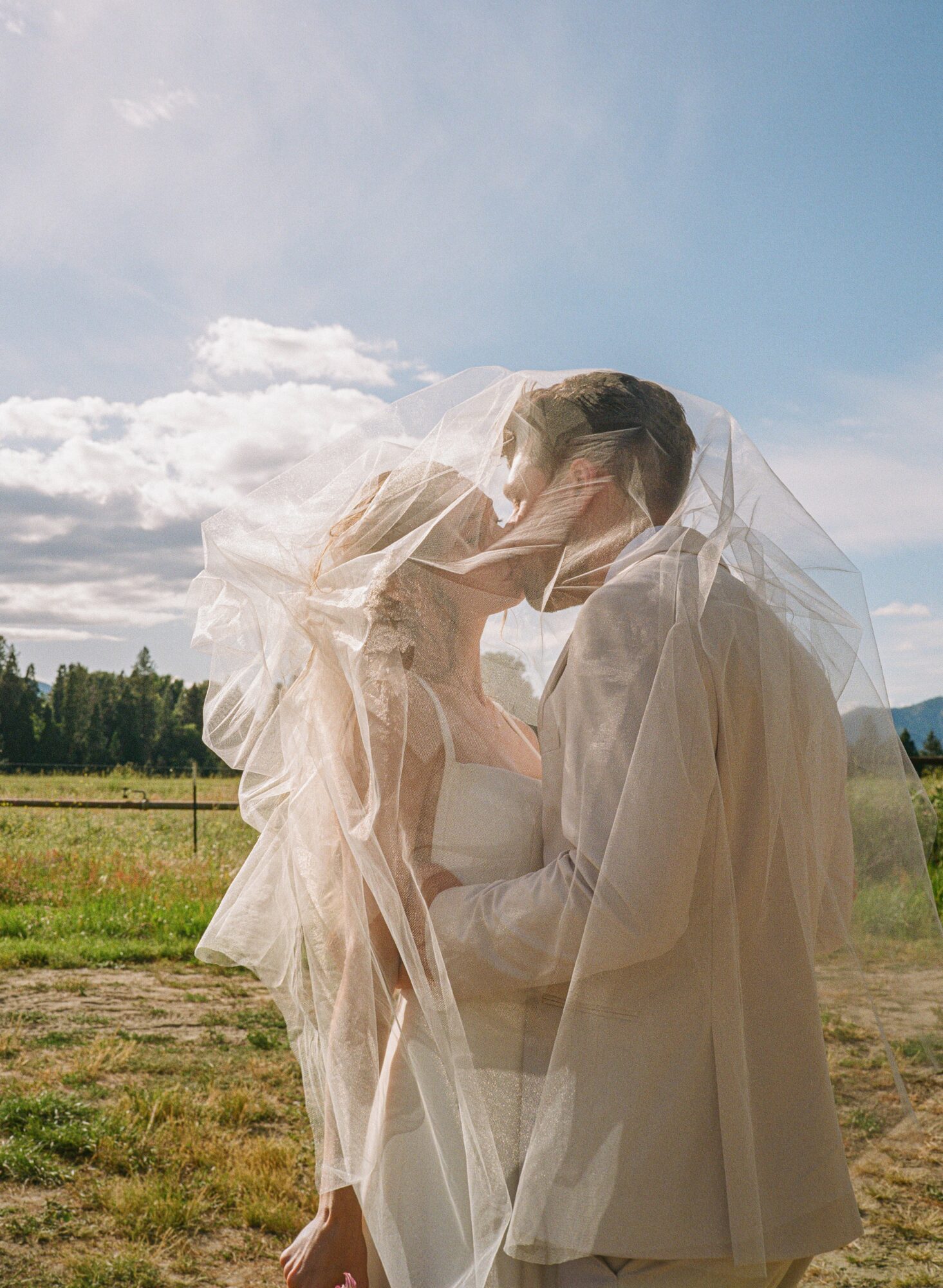 Two people, one with a veil, share an intimate moment outdoors with a field and sky in the background.