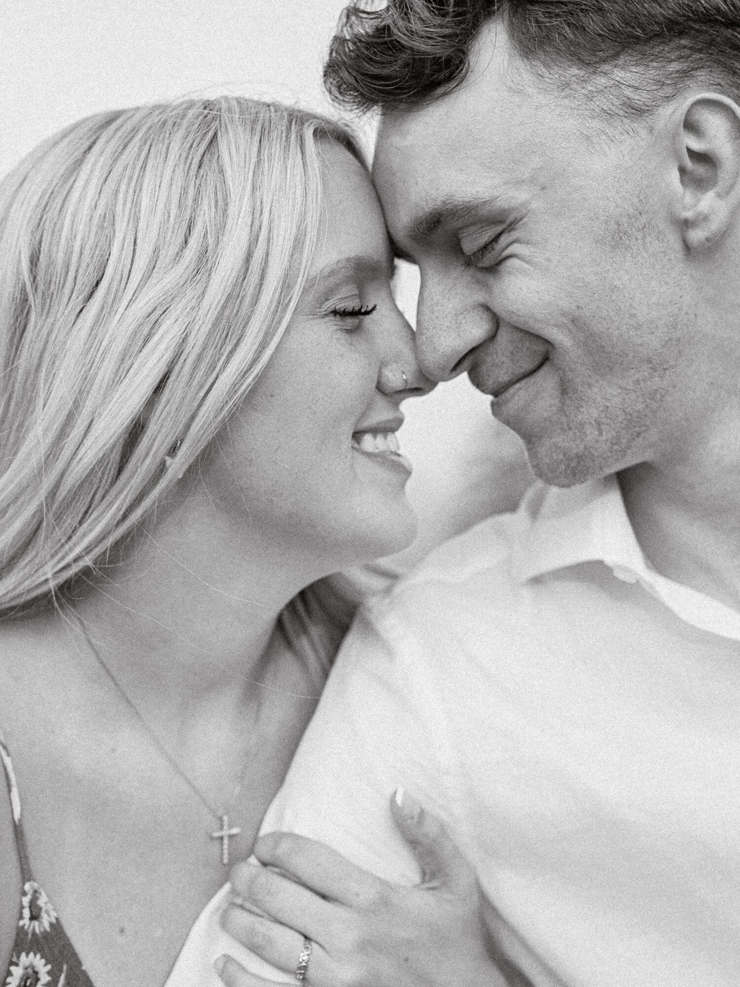 Close-up of a smiling couple with foreheads touching, woman wearing a cross necklace, black and white photo.