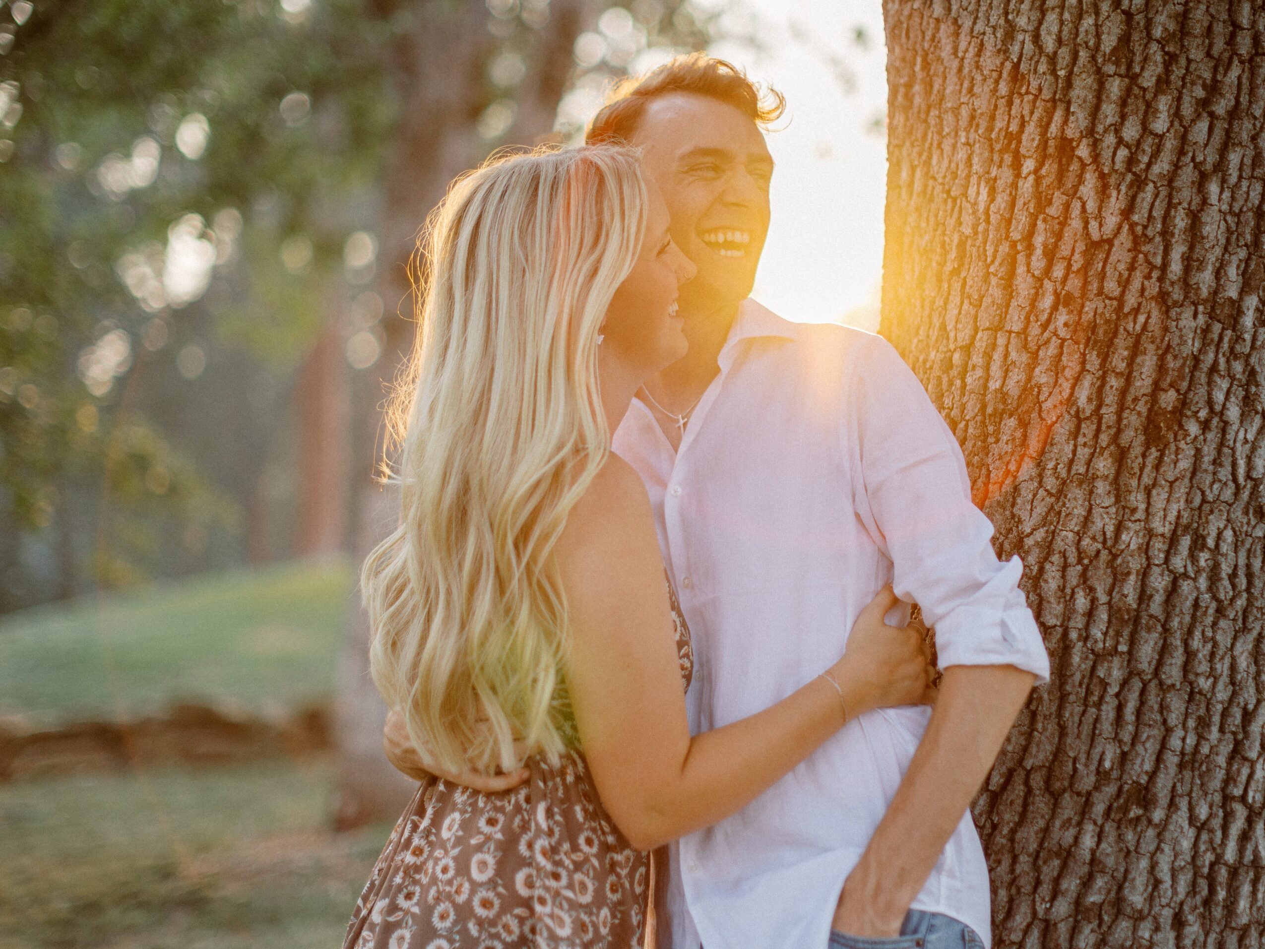 A smiling couple leans against a tree outdoors during sunset, with sunlight shining behind them.