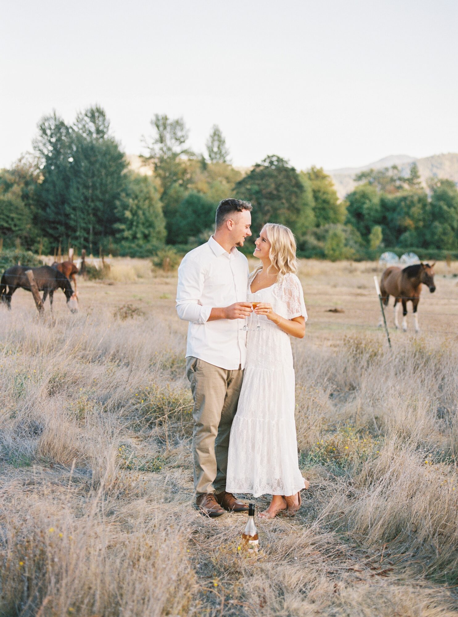 A couple stands close together in a field with horses, trees, and hills in the background, holding hands and looking at each other.