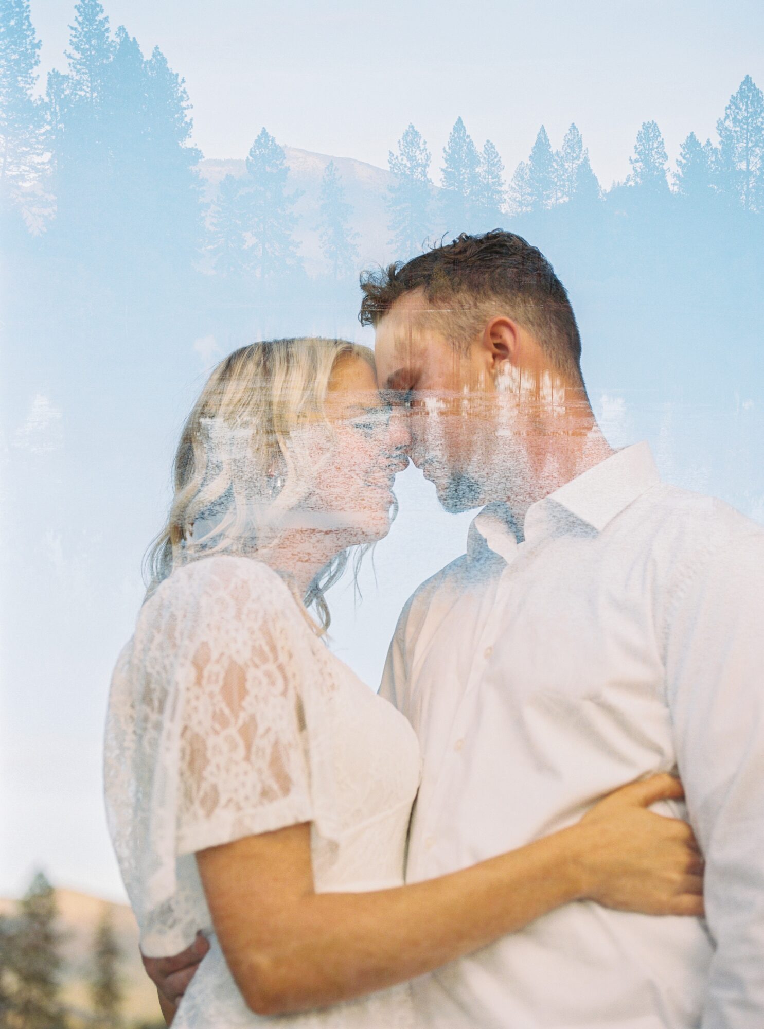 A couple with foreheads touching, smiling, outdoors with trees in the background, wearing light-colored clothing.
