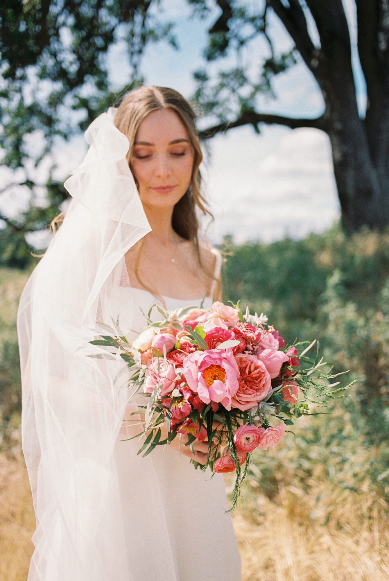 A bride with a veil holding a pink and white flower bouquet outdoors, trees and sky in background.