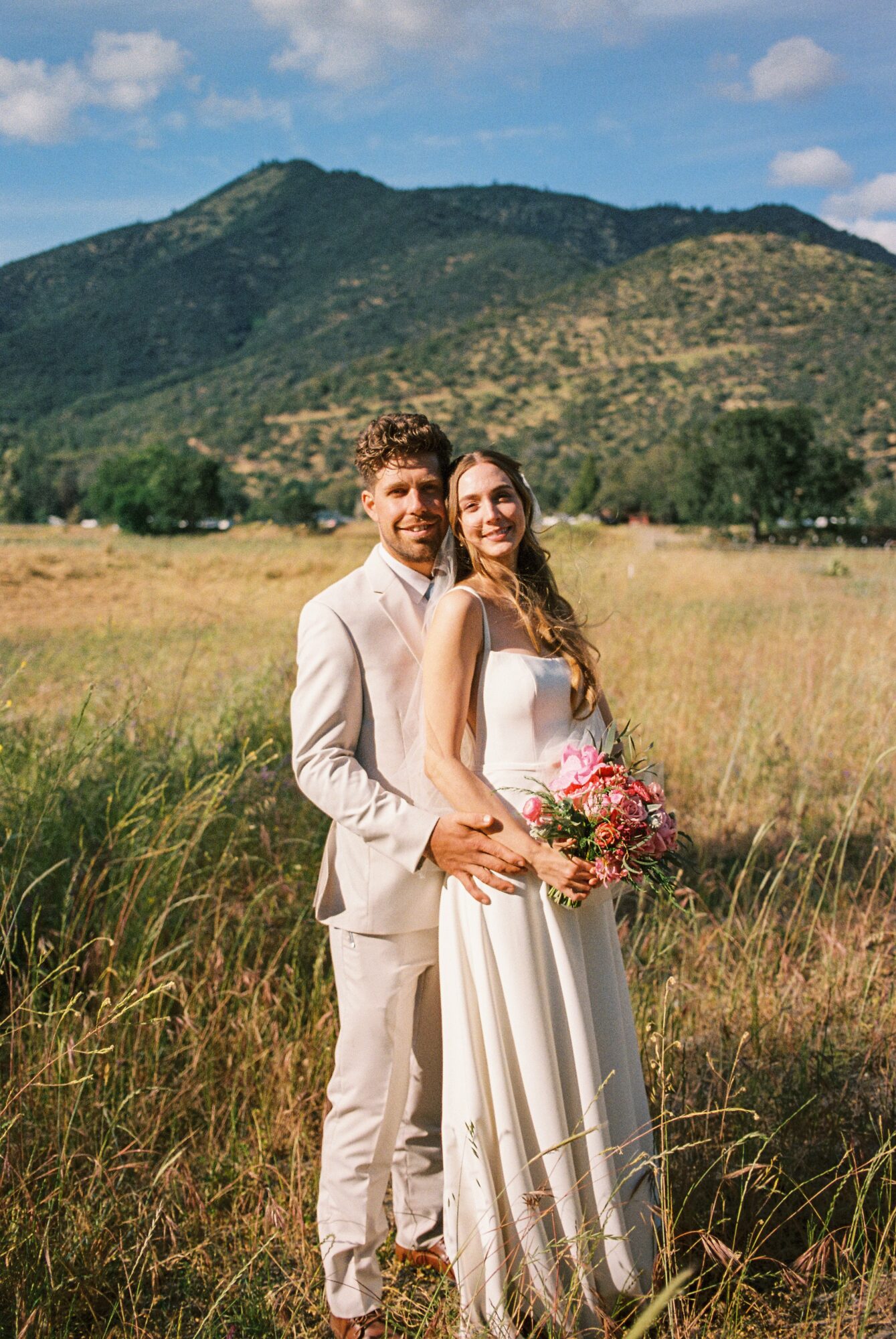A couple in wedding attire standing in a grassy field with mountains and blue sky in the background, holding a bouquet.