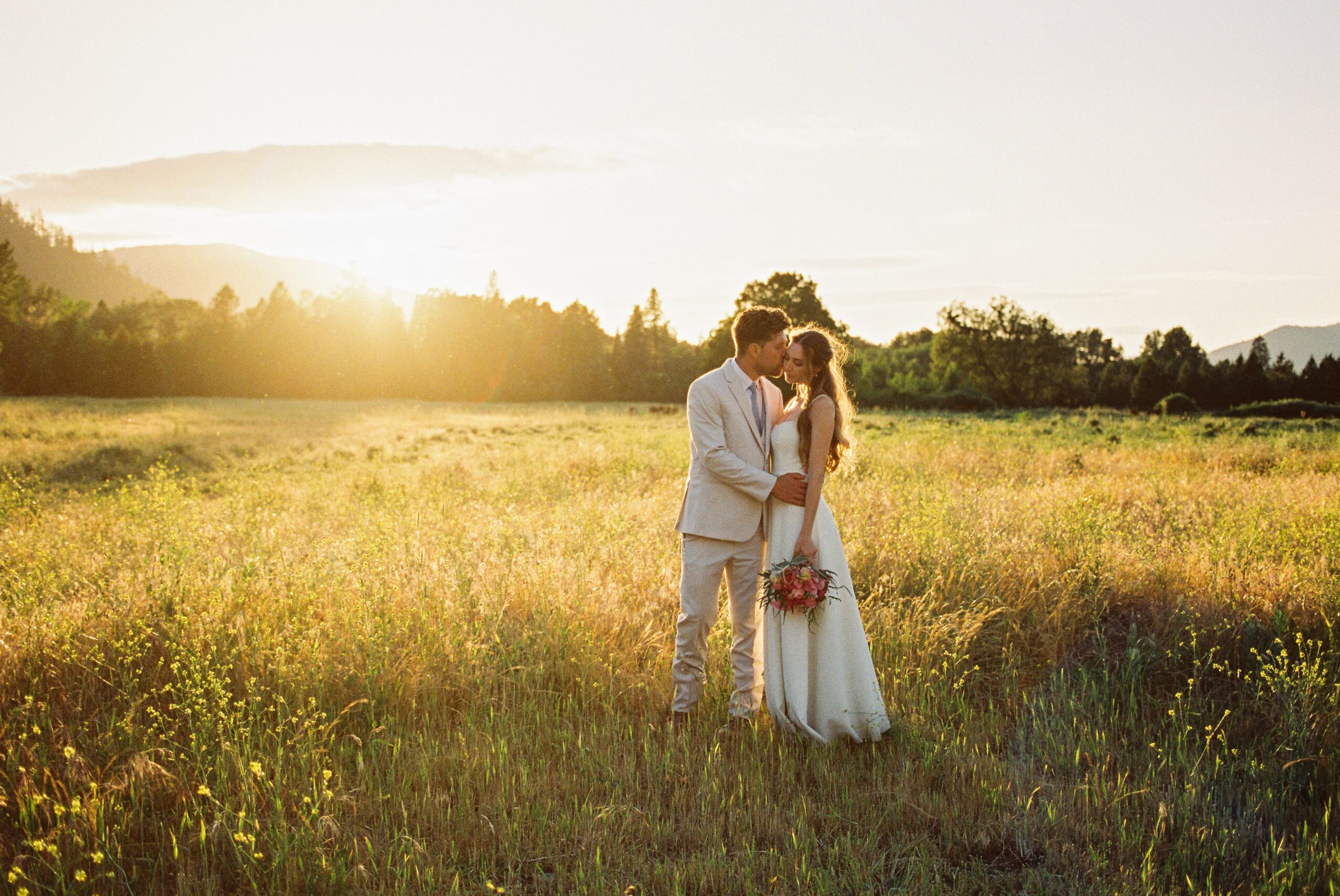 A couple in wedding attire stands in a field at sunset, embracing and holding a bouquet of flowers.