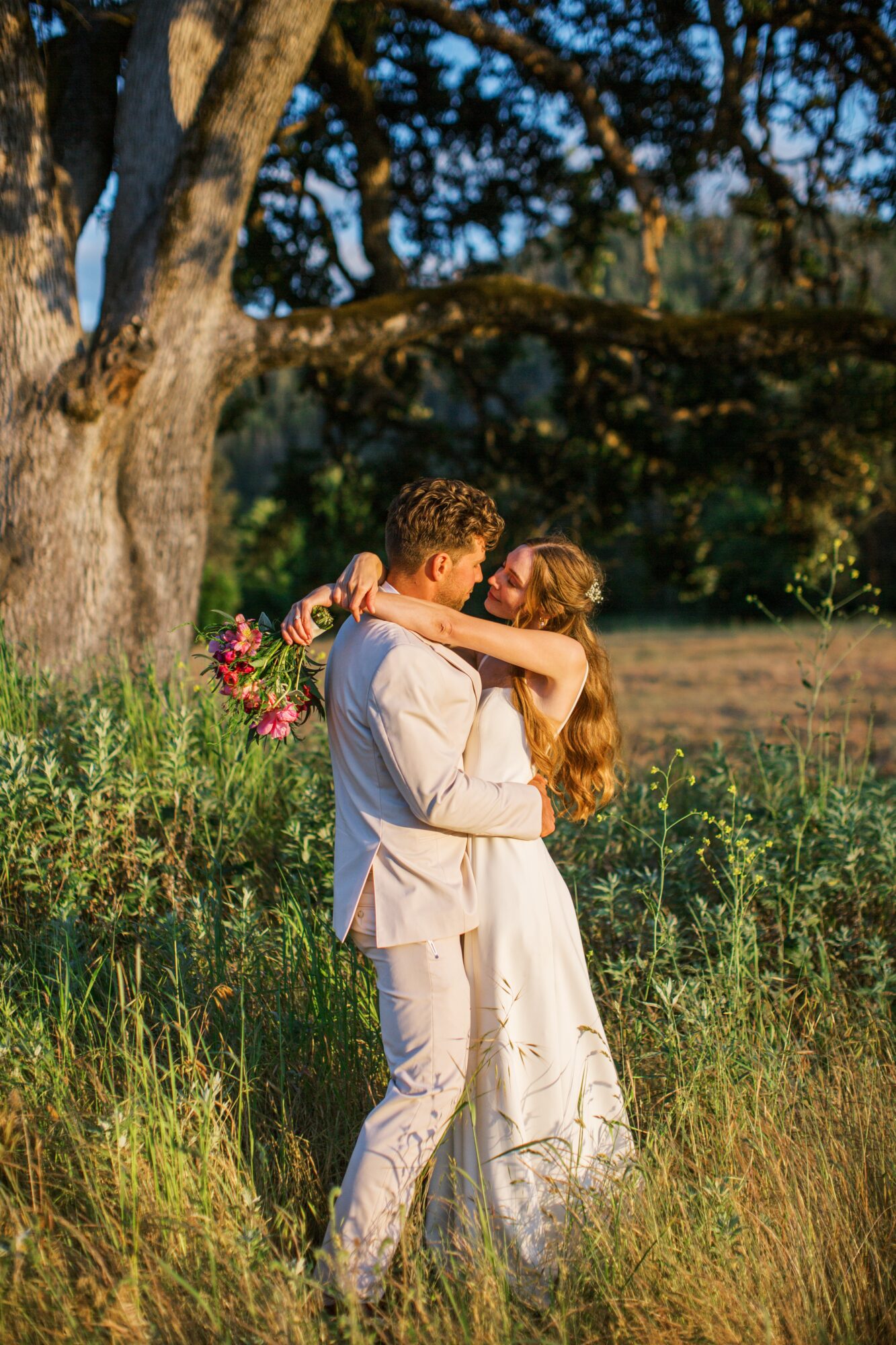 A couple embraces outdoors near a large tree, with sunlight and greenery in the background.