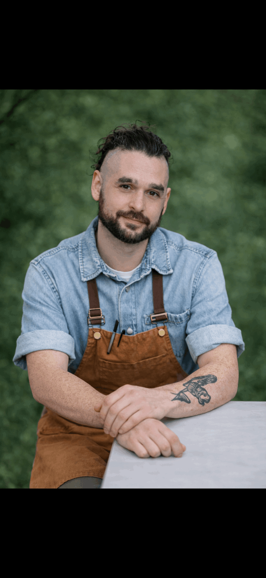 Man with beard and glasses wearing denim shirt and apron, sitting outdoors with greenery background.