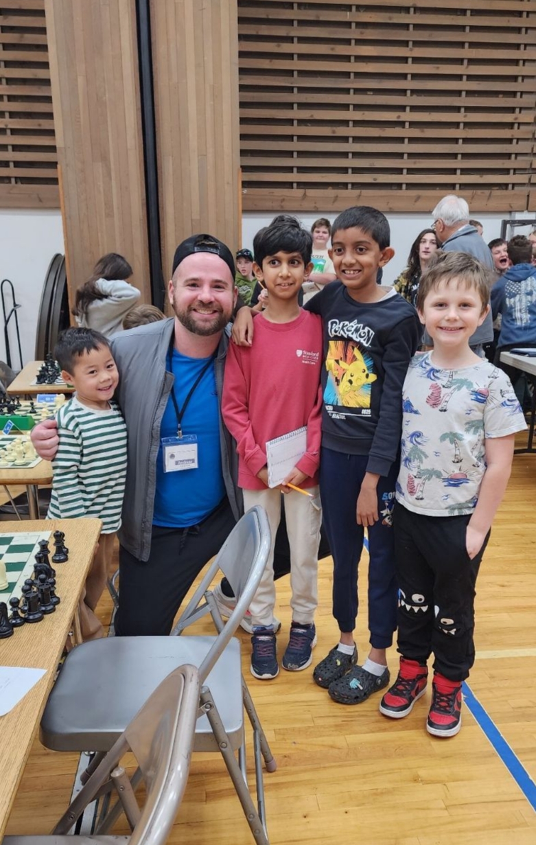 Man with four children in a room with wooden walls and tables, some children sitting at tables in the background.