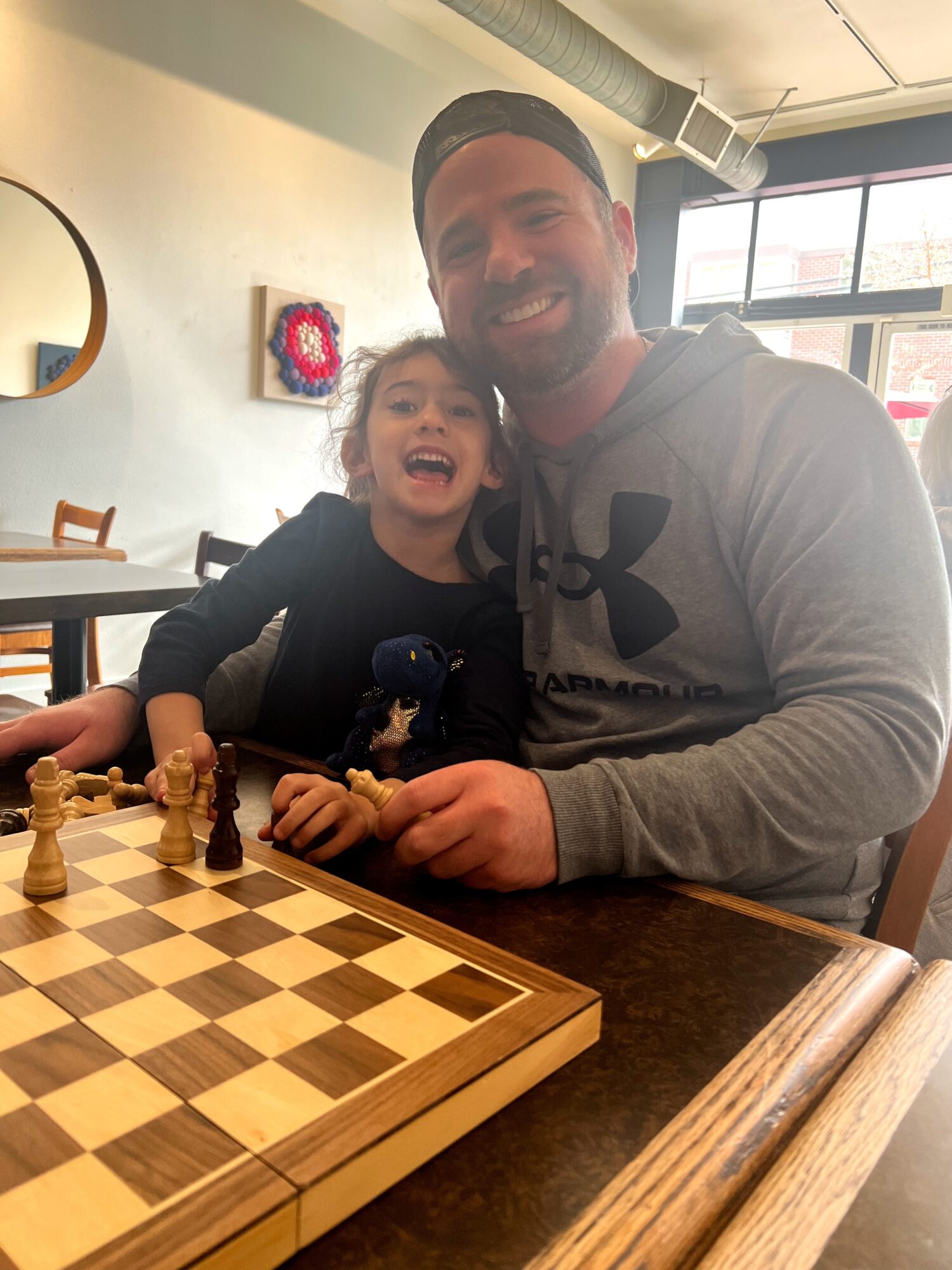 A man and a young girl sit at a table with a chessboard, smiling and laughing indoors with sunlight coming through windows.
