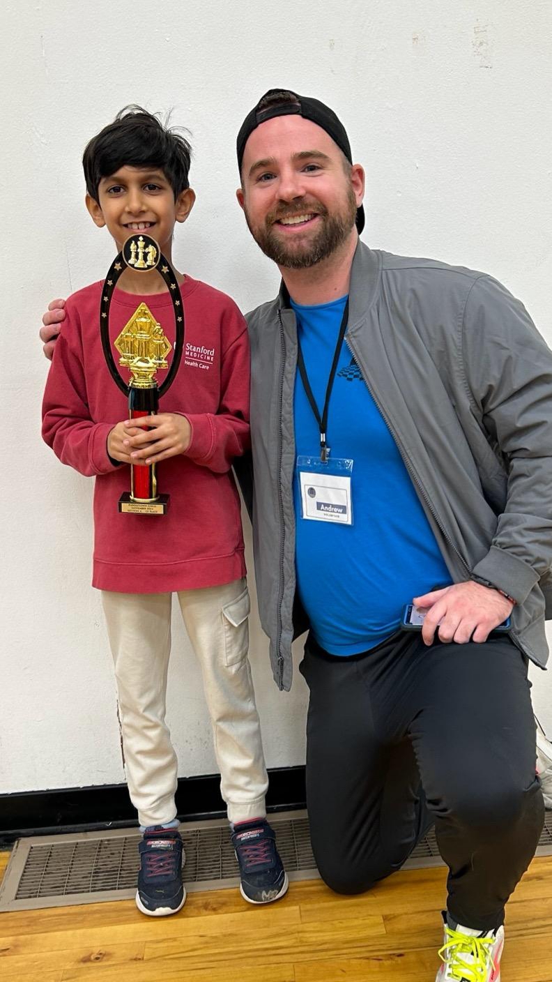 A young boy and a man pose together, smiling, with the boy holding a trophy. They are indoors against a plain wall.