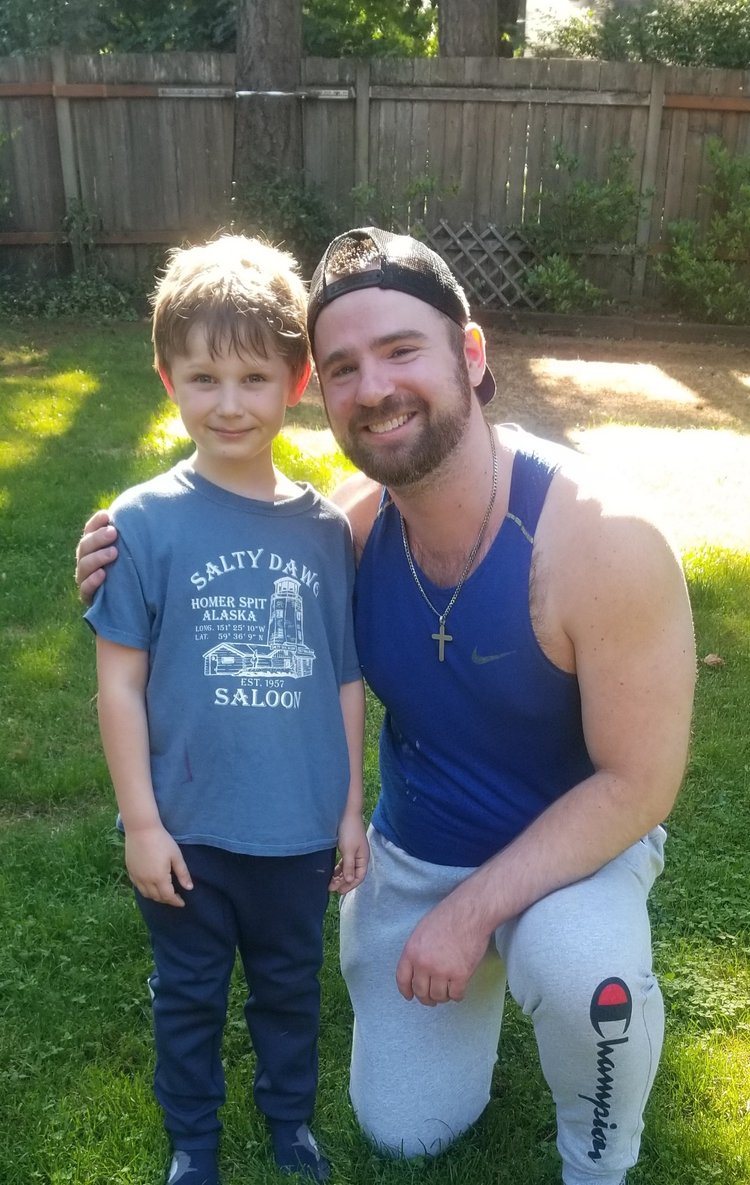A man and a young boy outdoors in a backyard, smiling, with a wooden fence and trees in the background.