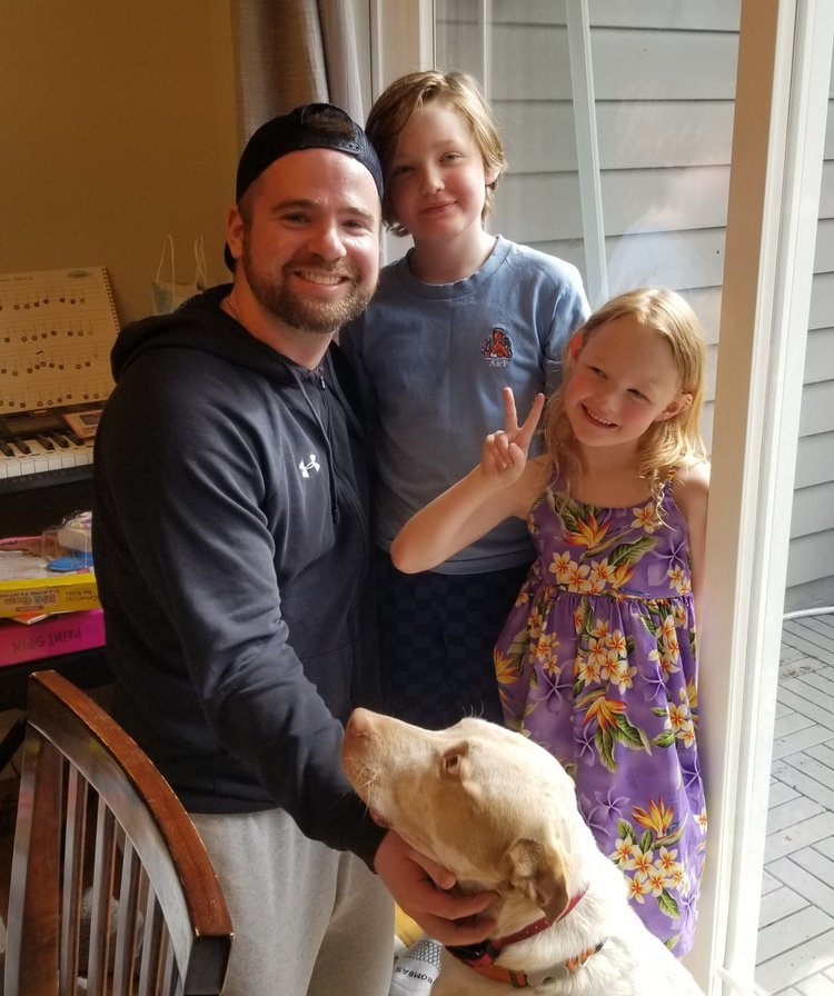 A man with two young girls and a dog indoors near a sliding glass door, smiling and making a peace sign.
