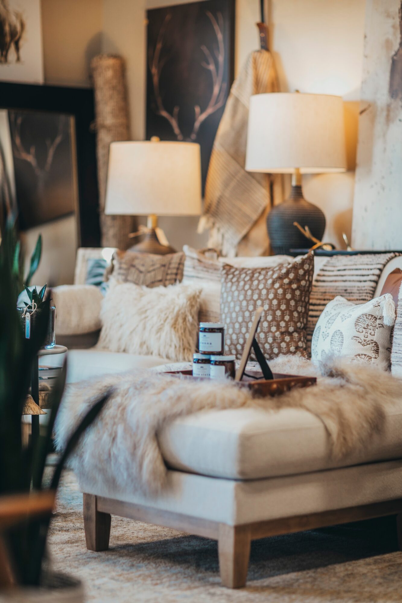 Cozy living room with two table lamps, cushions, and decorative items on a sofa, with artwork and plants in the background.