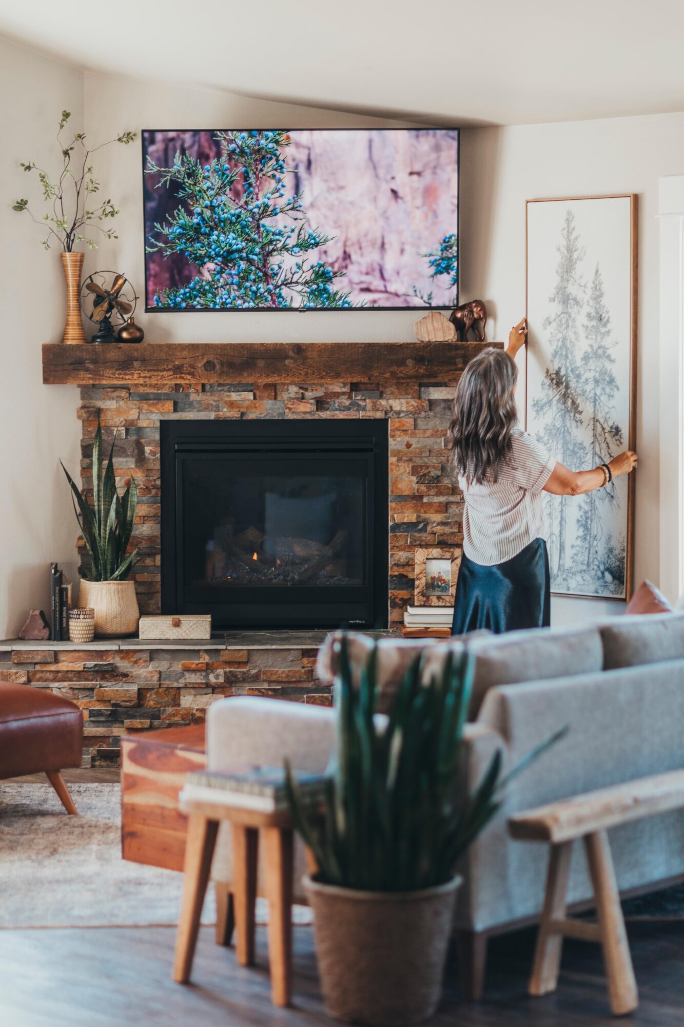 Person cleaning a large framed artwork on a wall in a cozy living room with a brick fireplace and television above.