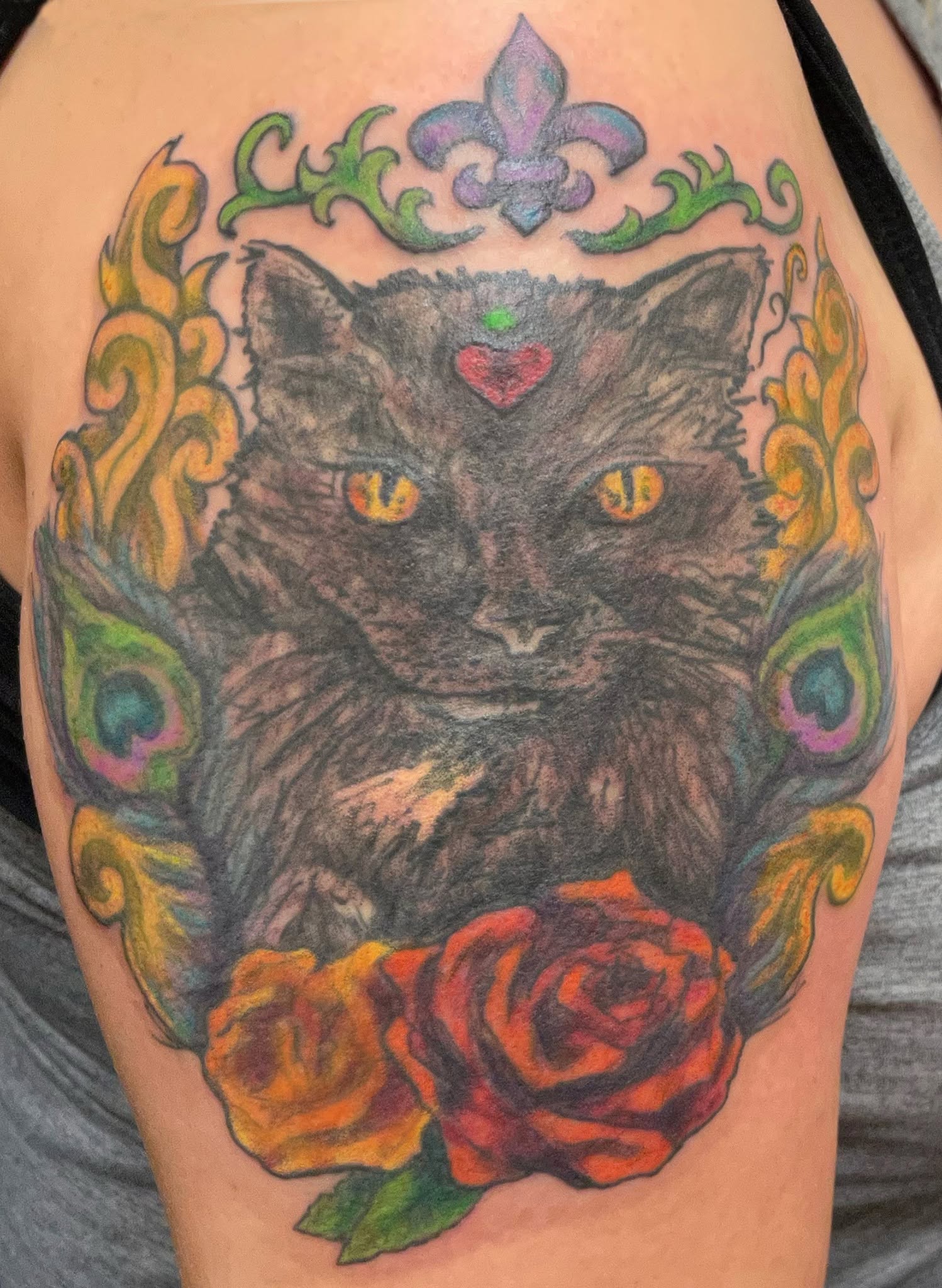 Tattoo of a black cat with yellow eyes, a red heart on its forehead, surrounded by colorful decorative elements and roses.