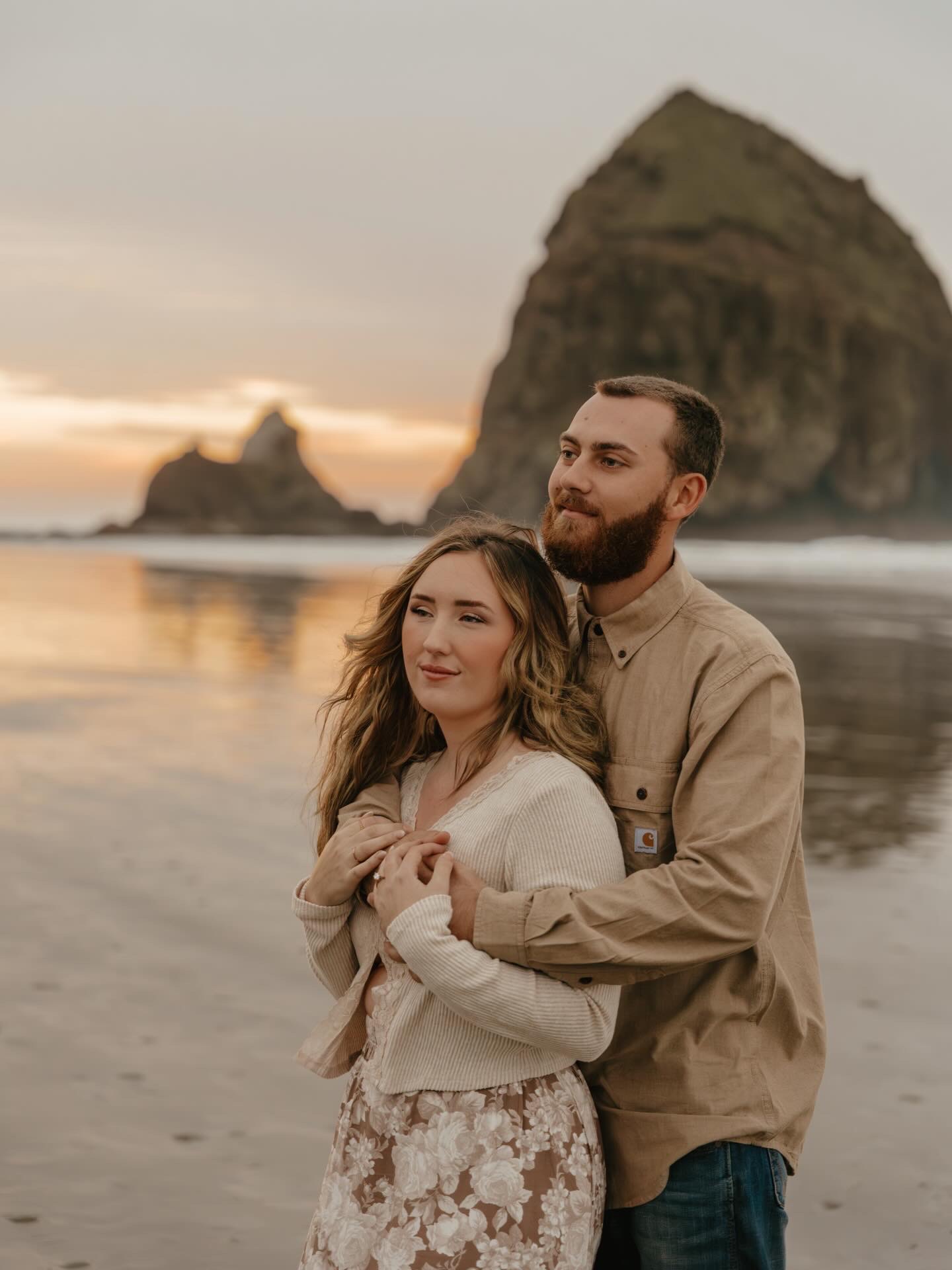A couple stands on a beach at sunset, with a large rock formation in the background, embracing each other.