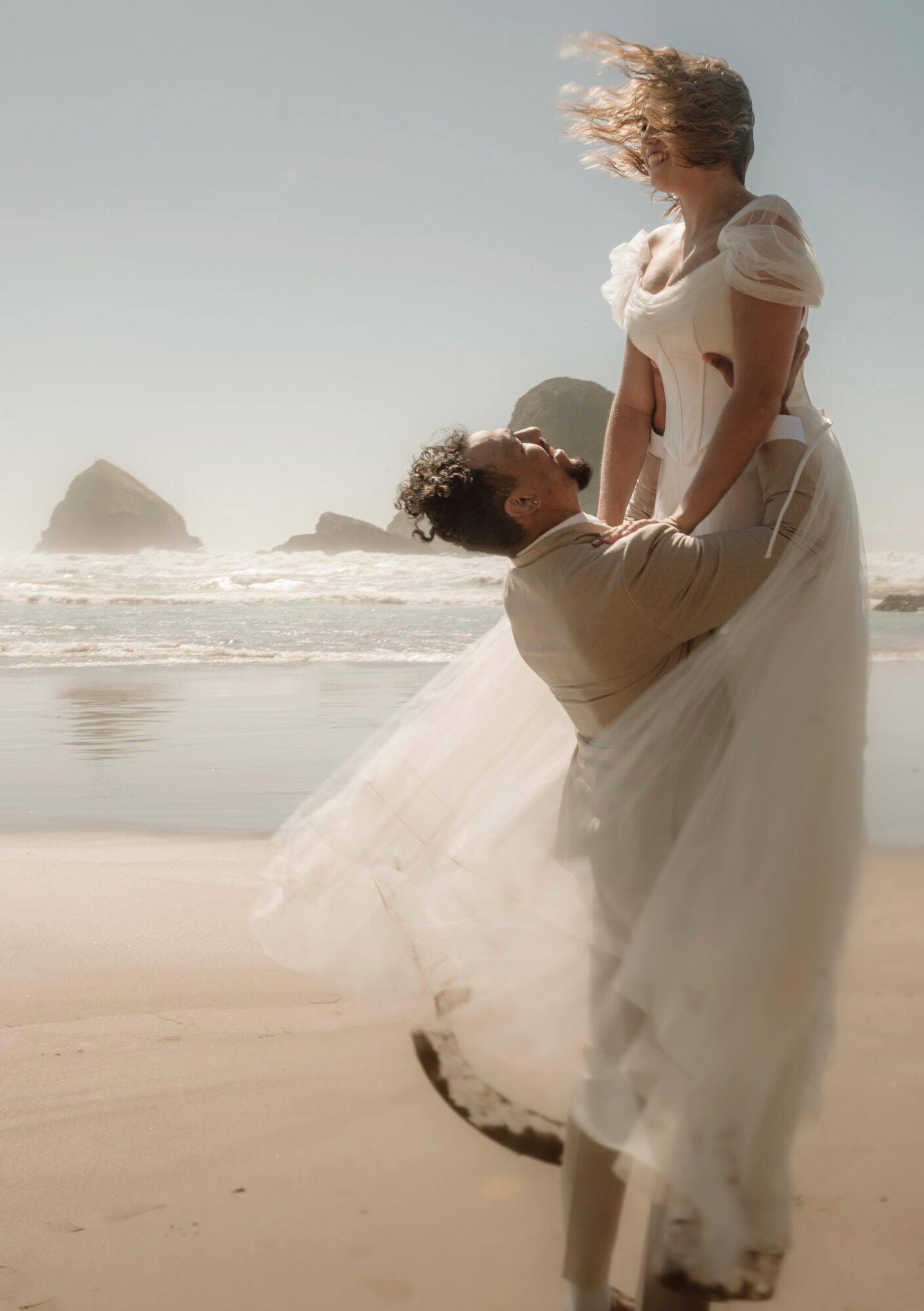 A couple on the beach, with the man lifting the woman, her hair blowing in the wind, ocean waves in background.