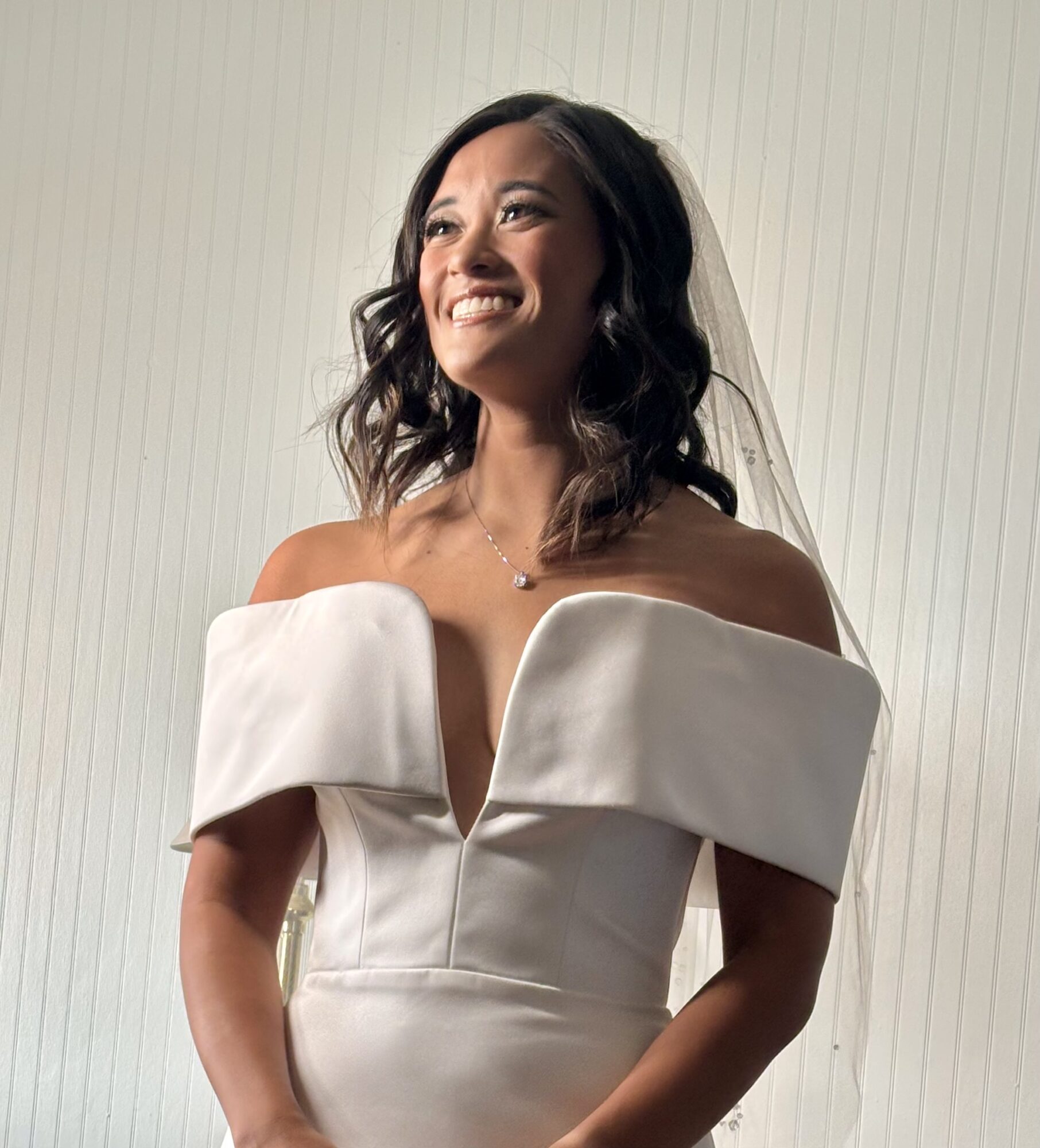 Woman in wedding dress smiling, off-shoulder gown with large collar, dark wavy hair, standing against a light background.