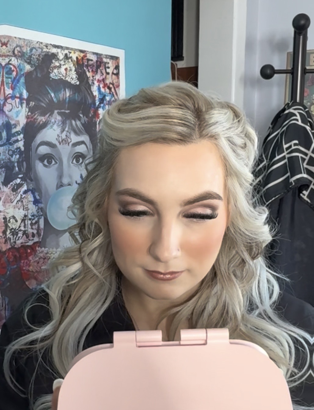 Woman with long, wavy blonde hair and makeup, looking down, in front of colorful artwork, indoors.