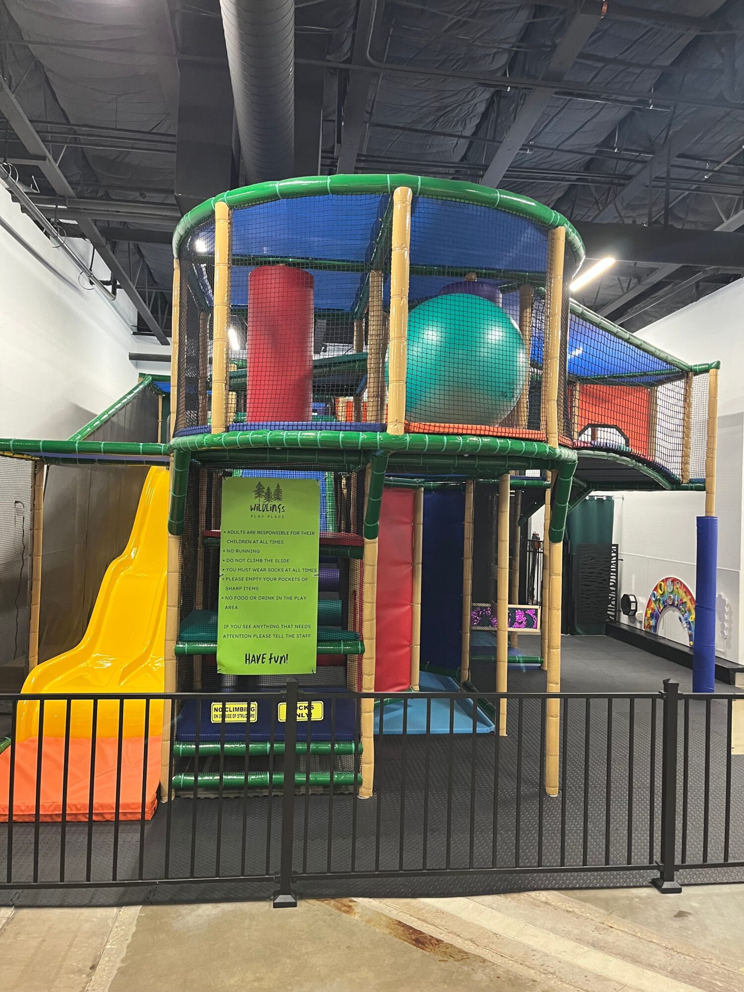 Indoor play area with a green and blue structure, slide, and large ball, surrounded by black fencing.