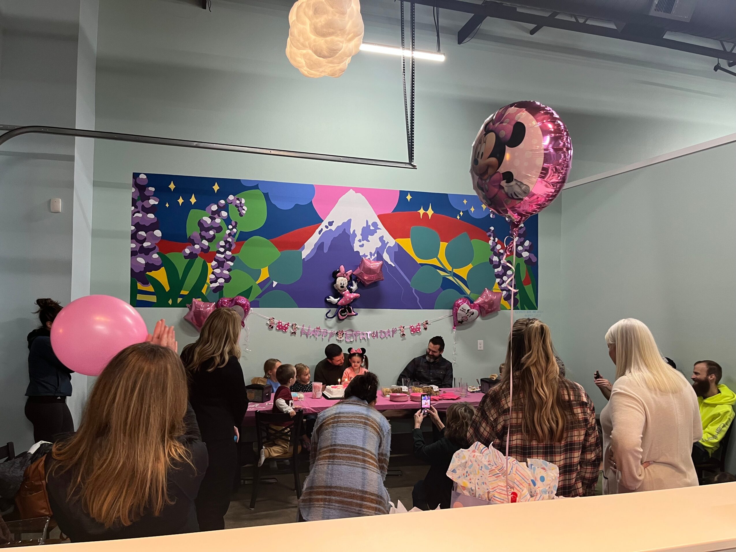 People seated at a decorated party with balloons and a colorful backdrop featuring a mountain and cartoon characters.