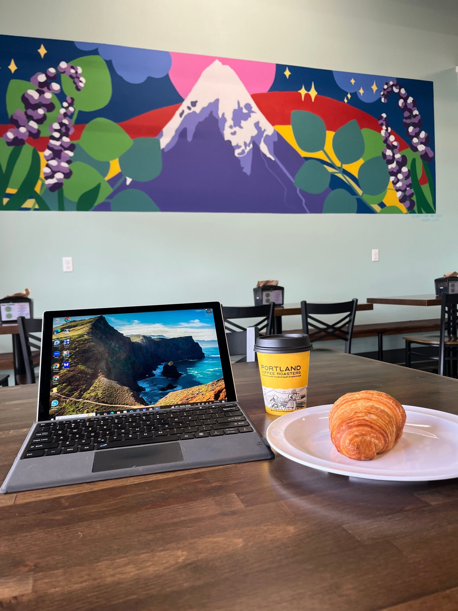 Laptop on wooden table with a mountain landscape wallpaper, a plate with a pastry, and a bottle, in a room with a colorful mural.
