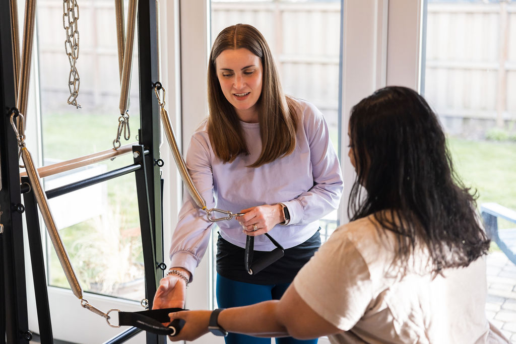 Two women in a room with large windows, one holding a tool and the other observing, engaged in a task.