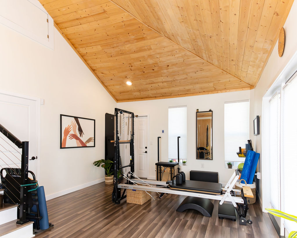 Pilates reformer machine in a bright room with wooden ceiling, white walls, and large windows, with a framed artwork on the wall.
