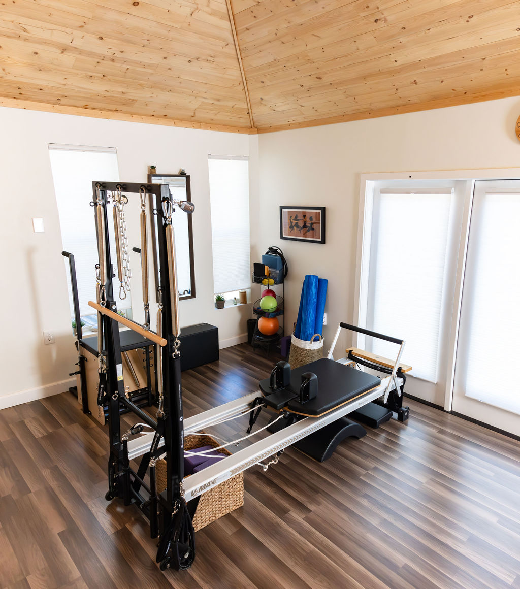 Pilates reformer machine in a bright room with wooden ceiling and floor, windows, and exercise equipment nearby.