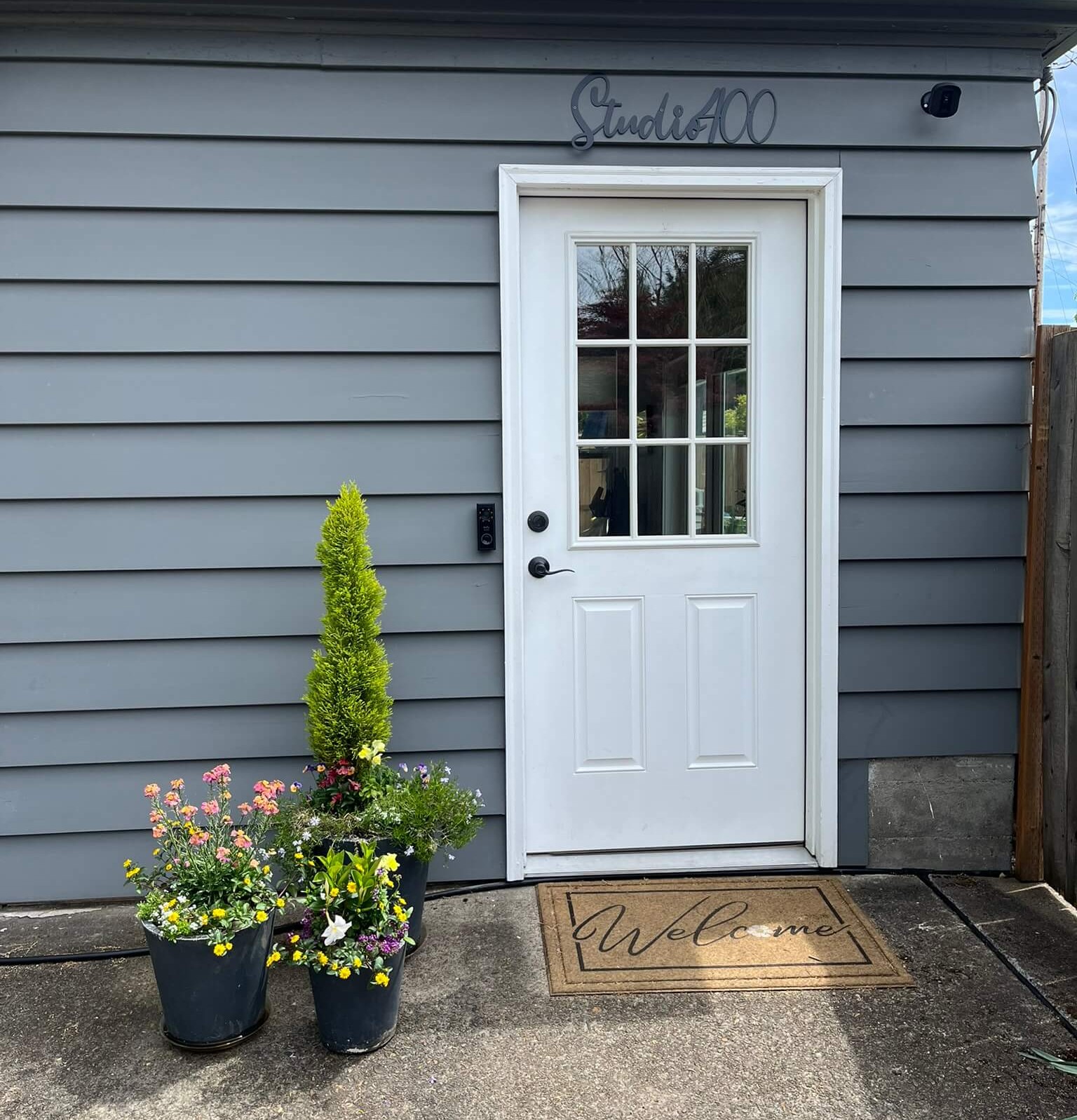 White door with window panes on a gray house exterior, potted flowers and a welcome mat outside.