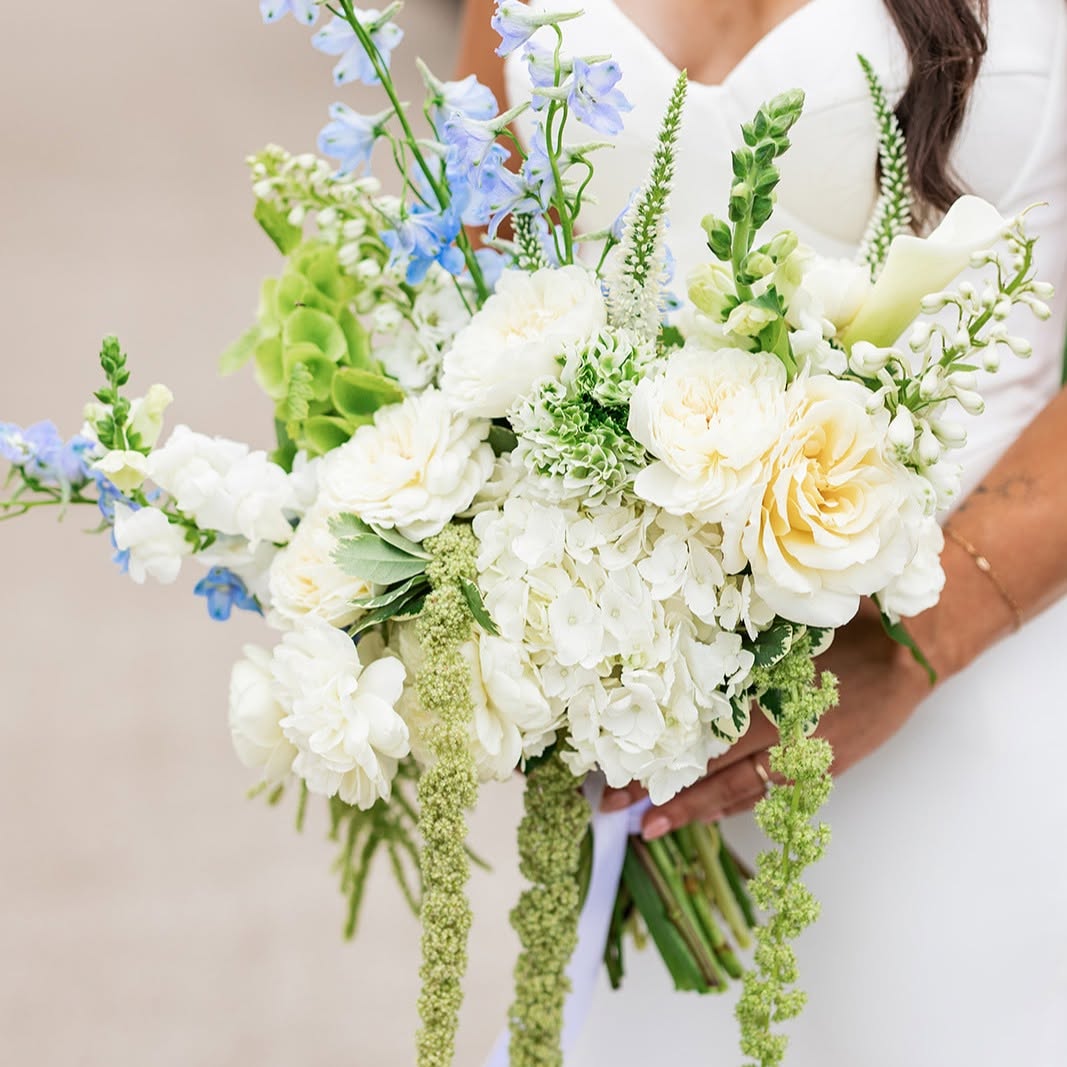 Person holding a large bouquet of white and light green flowers with some blue accents, wearing a white dress.