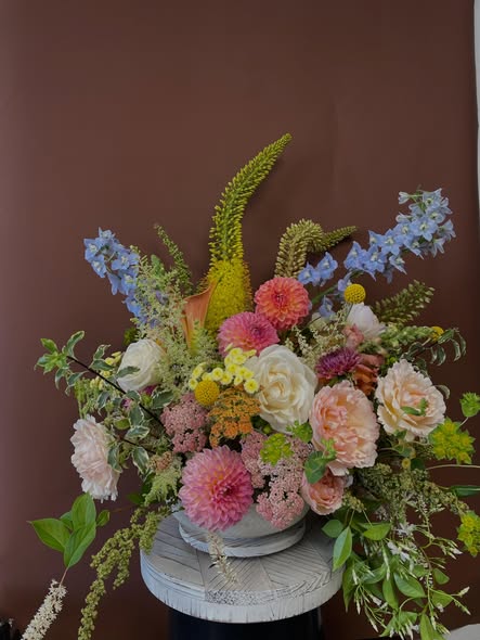 Colorful flower arrangement with pink, white, yellow, and green flowers in a white vase on a small round table against a brown background.