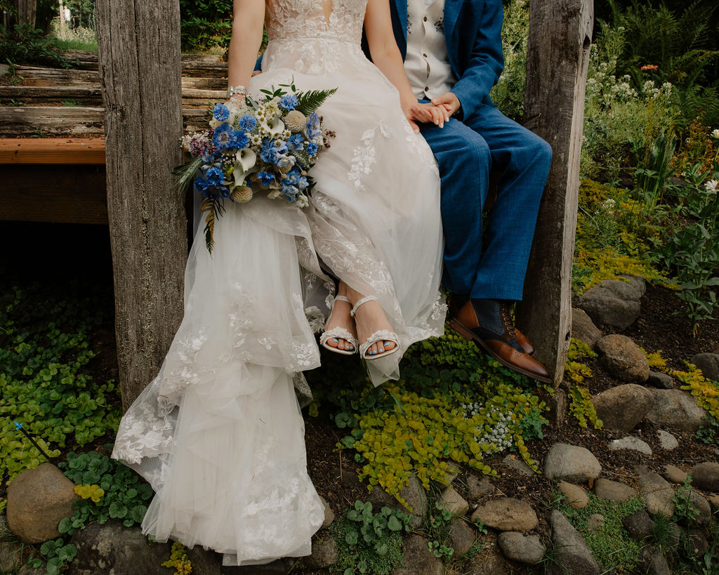 Bride and groom sitting on a wooden bench outdoors, bride holding a bouquet, surrounded by greenery and rocks.