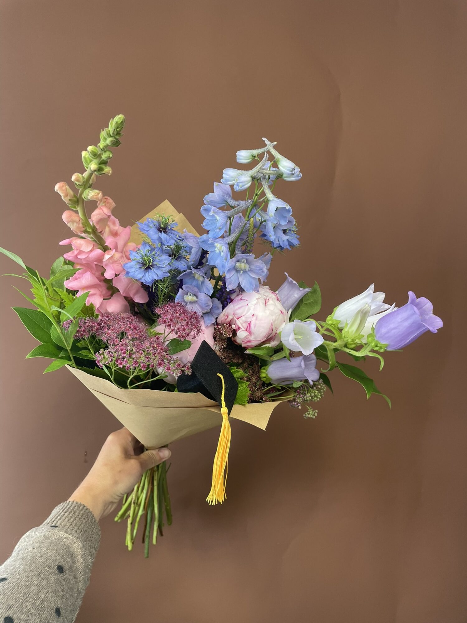 Person holding a colorful flower bouquet with pink, blue, and white flowers against a plain brown background.