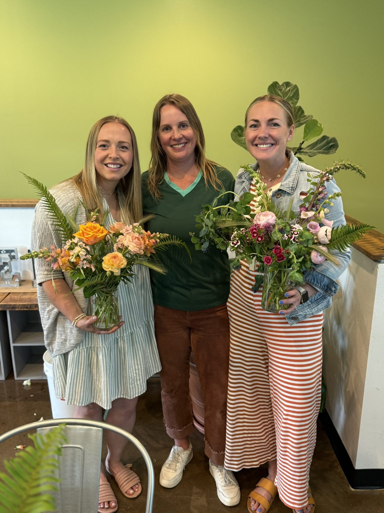 Three women smiling and holding bouquets of flowers indoors, with a green wall background.
