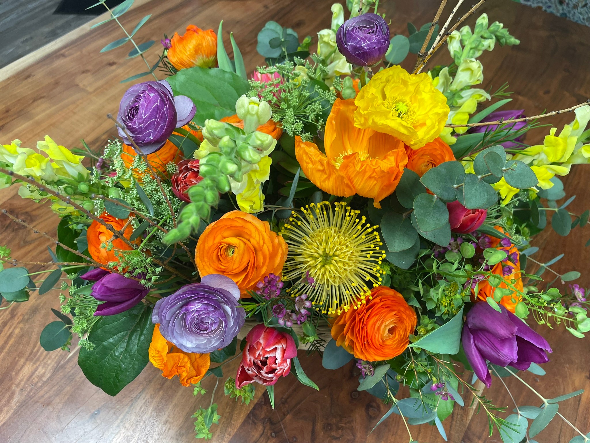 Colorful flower arrangement with purple, yellow, orange, and pink flowers, and green foliage in a vase on a wooden surface.