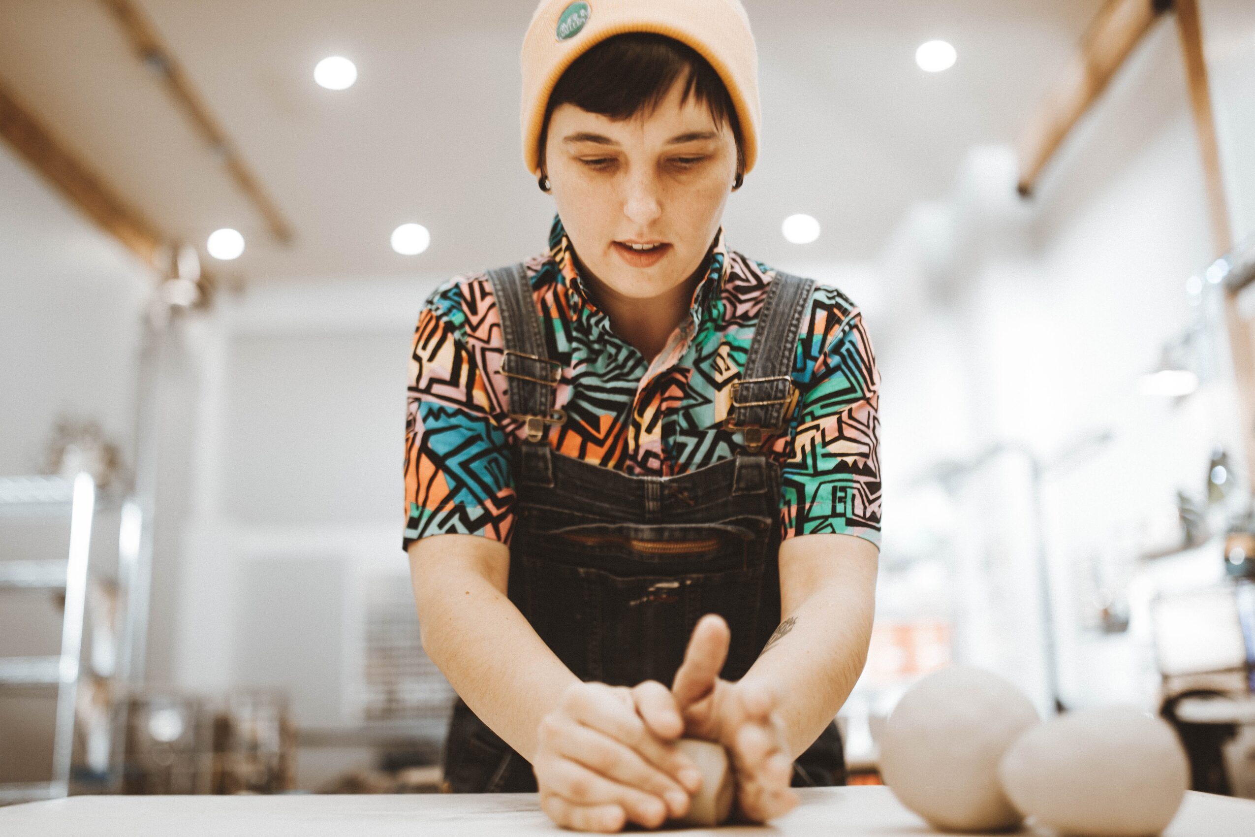 Person with short hair wearing a patterned shirt and white hat, working with dough in a kitchen, focused on hands.