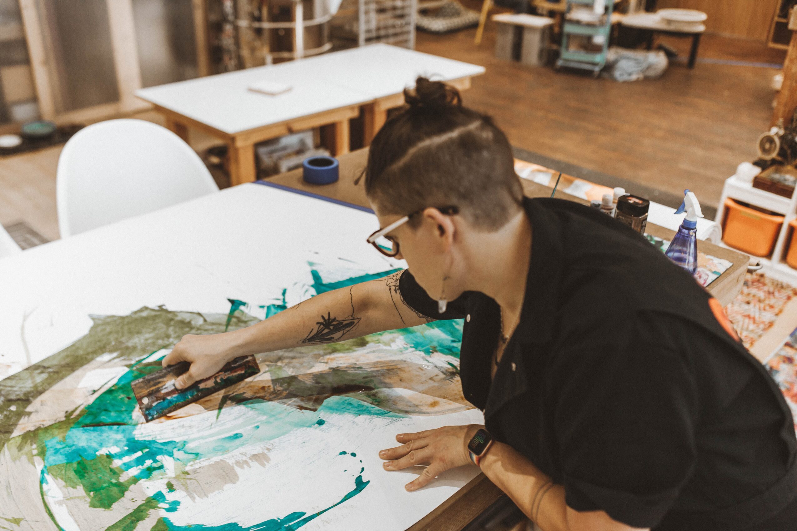 Person with short hair and glasses painting on a large canvas in a room with tables and shelves, using brushes and paints.