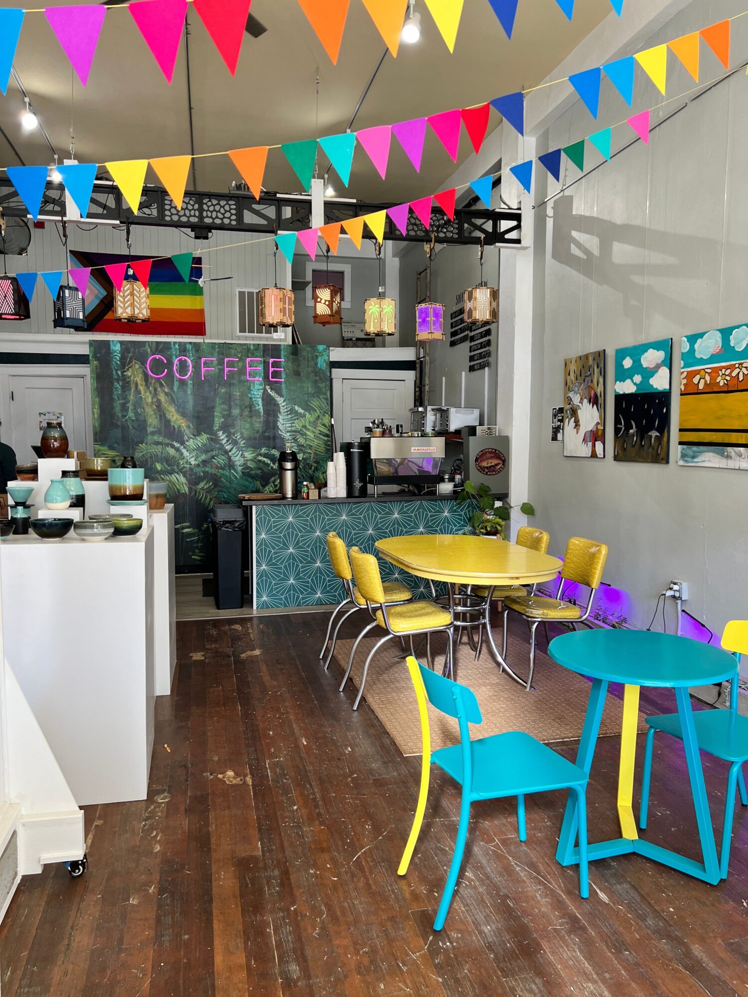 Colorful cafe interior with yellow and blue tables and chairs, decorated with triangular banners, and a mural with the word 'coffee'.