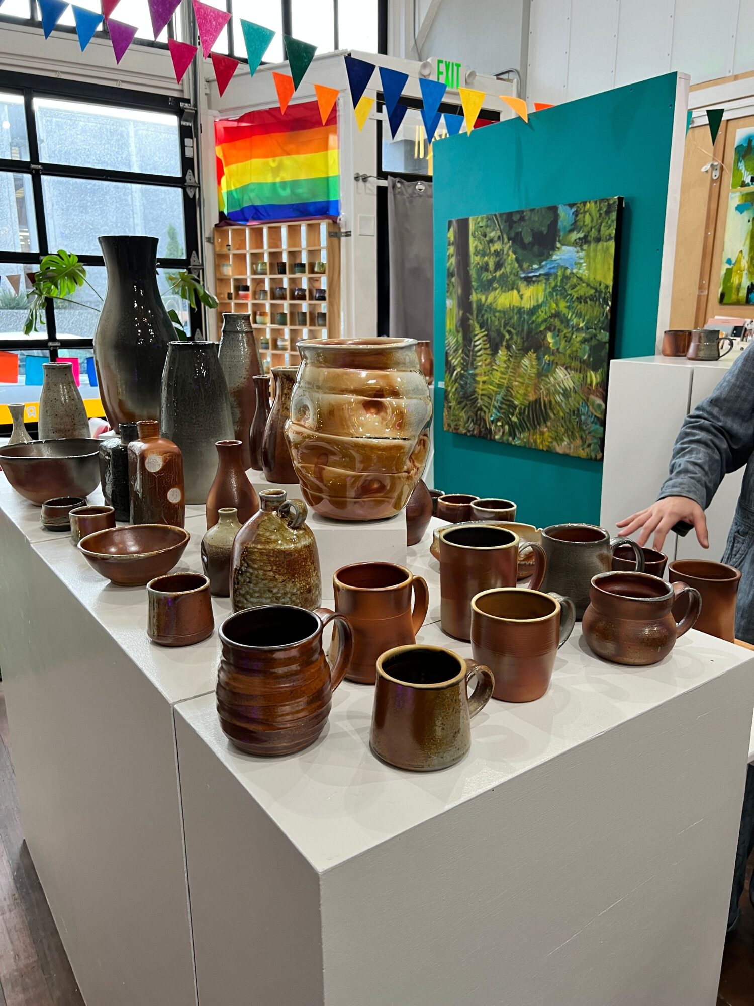 Display of various brown ceramic cups and a large glass jar on a white table, with a colorful rainbow flag and a window in the background.