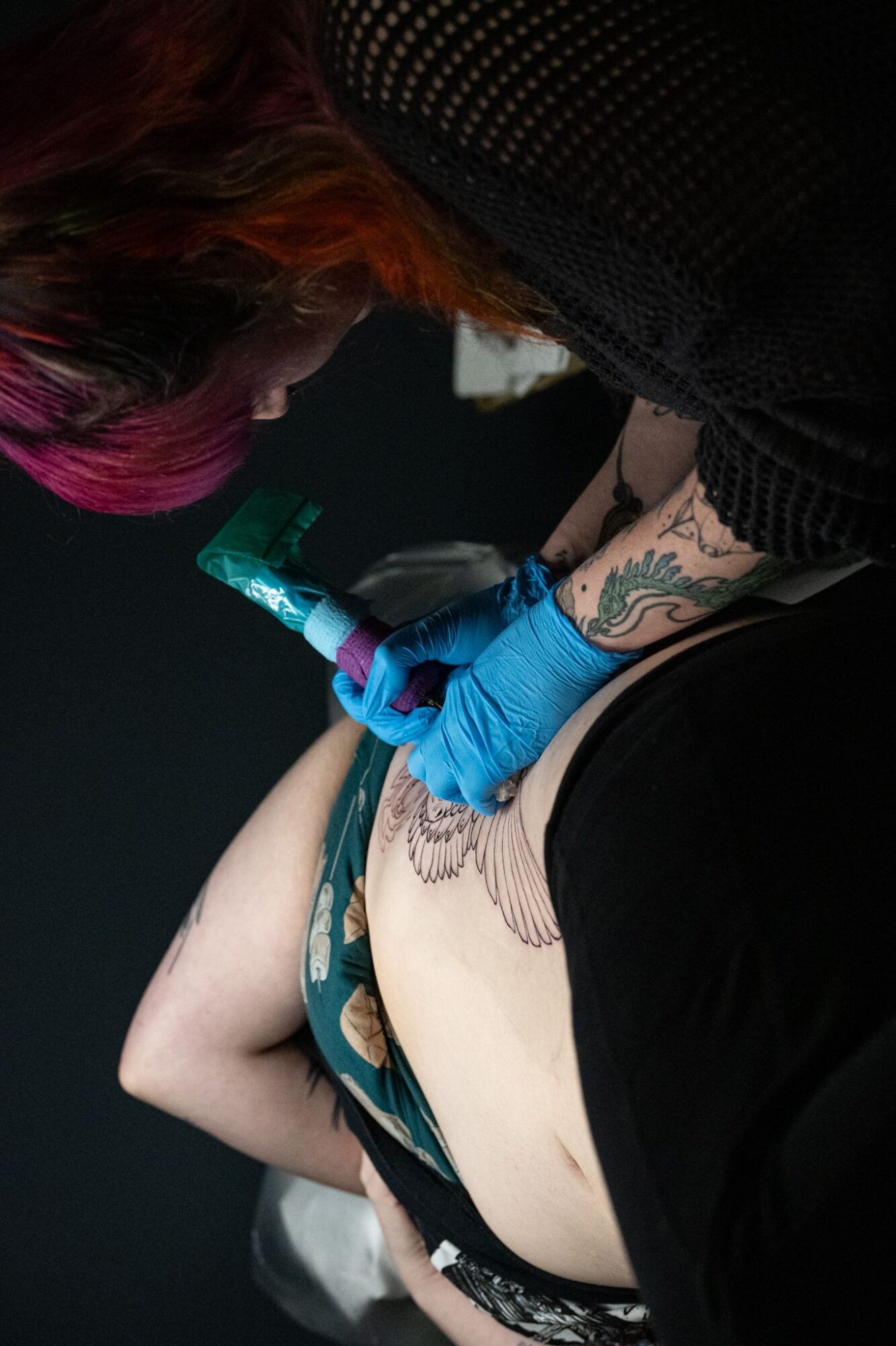Tattoo artist wearing blue gloves tattooing a person's side with a tattoo machine, dark background.