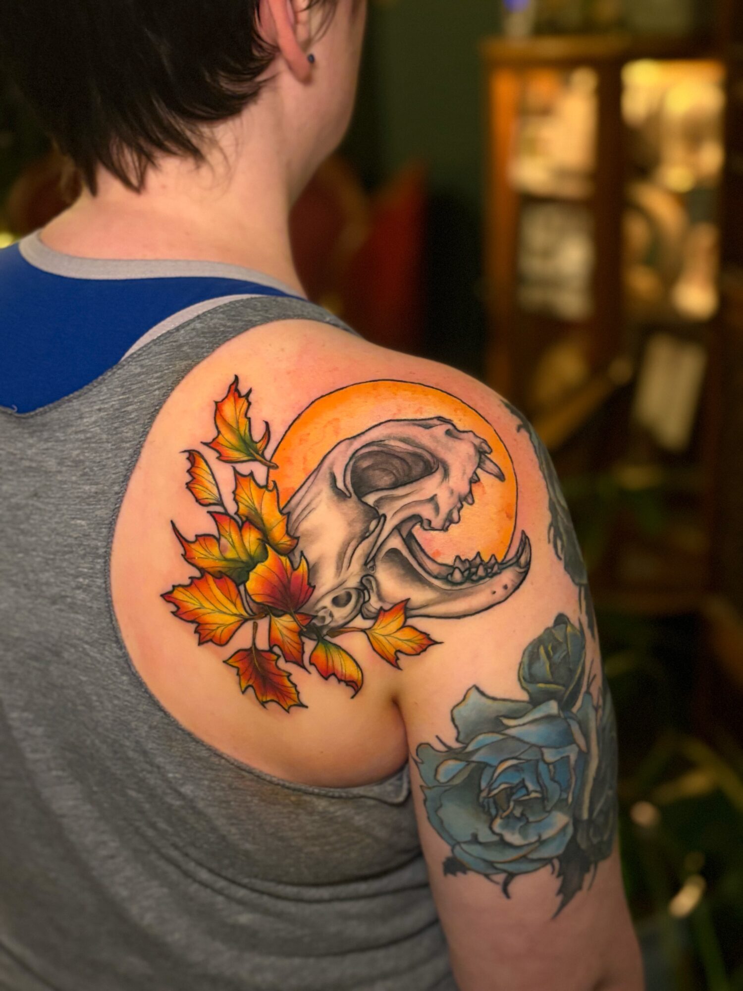 Tattoo of a skull with a large moon and autumn leaves on a person's shoulder, with a blue flower on the arm.