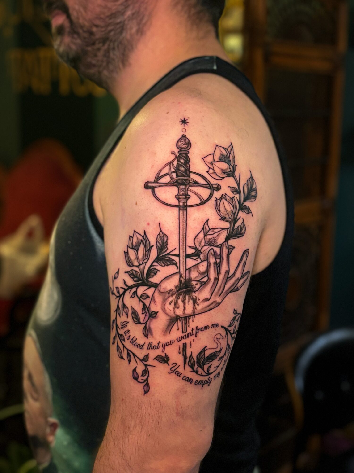 Tattoo of a sword with roses and a quote on a person's upper arm, with visible shoulder and part of chest.