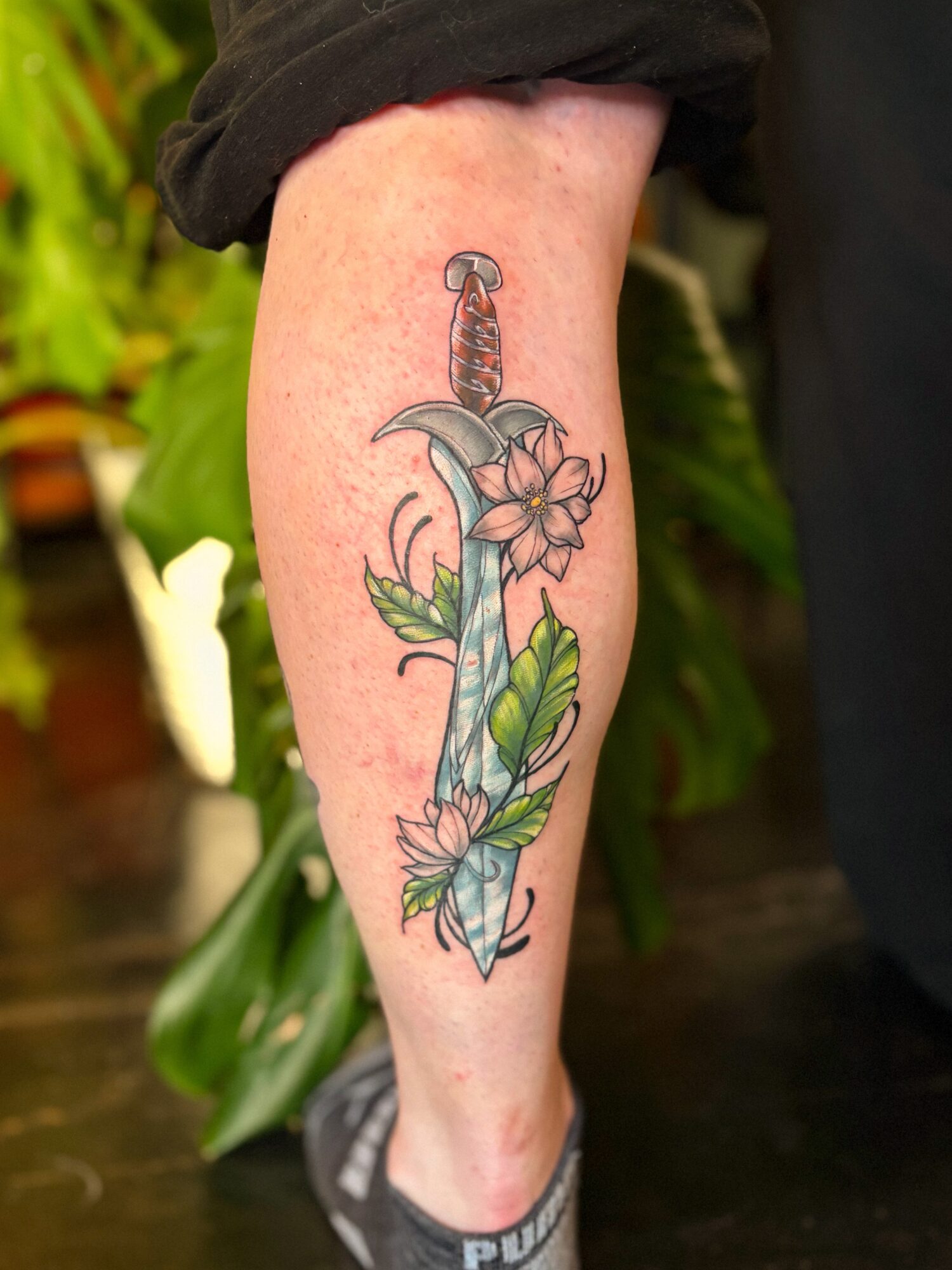 Tattoo of a dagger with flowers and leaves on a person's forearm, with a black sleeve visible at the top.