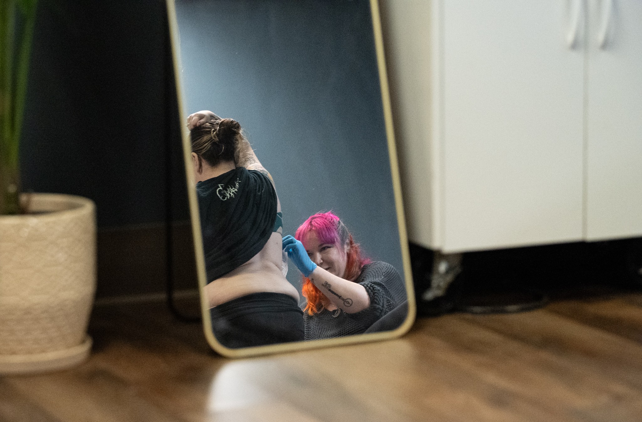 Child with pink hair and gloves reflected in a mirror, with another person nearby, on a wooden floor.