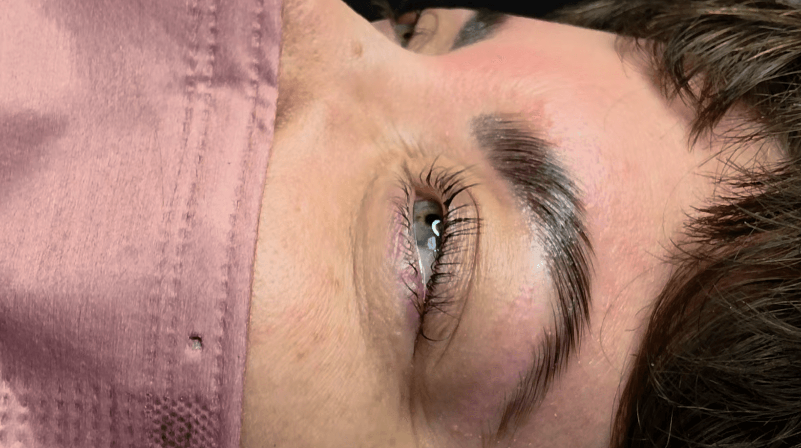 Close-up of a person's eye and eyebrow, with part of a pink face mask visible, lying sideways.