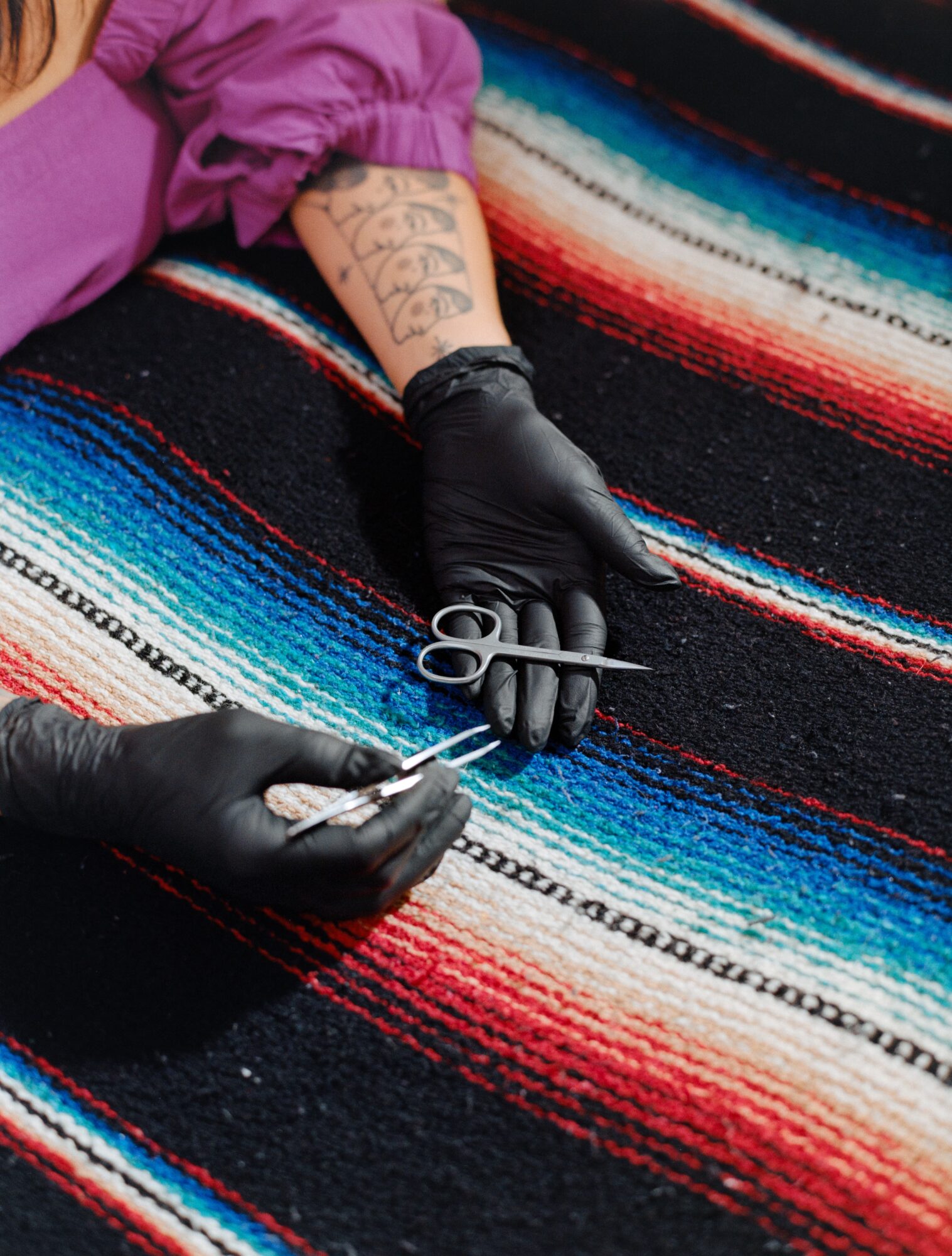 Person with tattooed arm and black gloves holding scissors and a needle, working on a colorful woven textile.